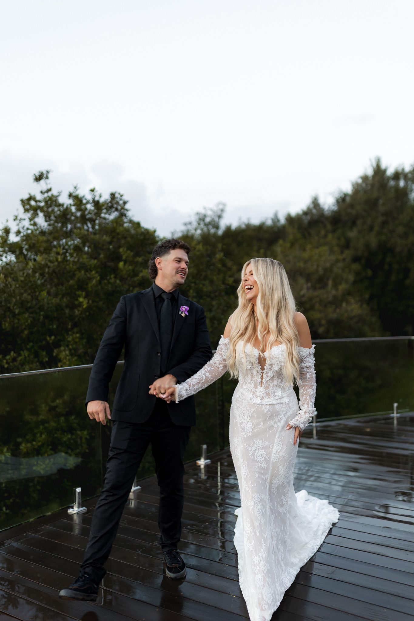 Bride in lace gown and groom in black suit hold hands laughing on a wet outdoor deck surrounded by greenery.
