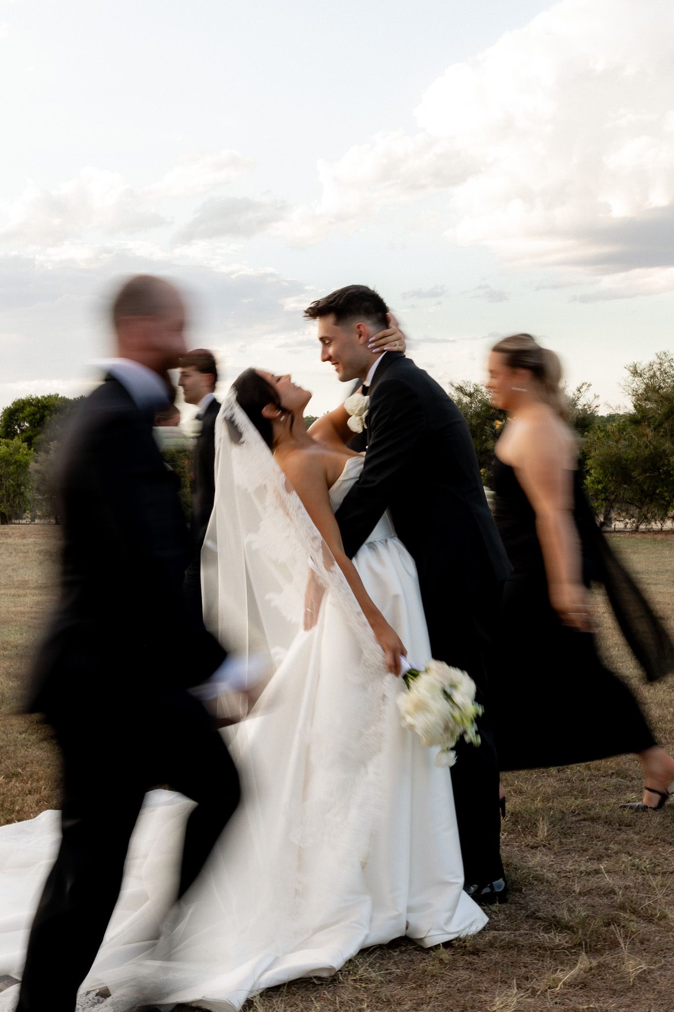 Bride and groom gazing at each other tenderly while wedding party walks past in artistic motion blur outdoors.