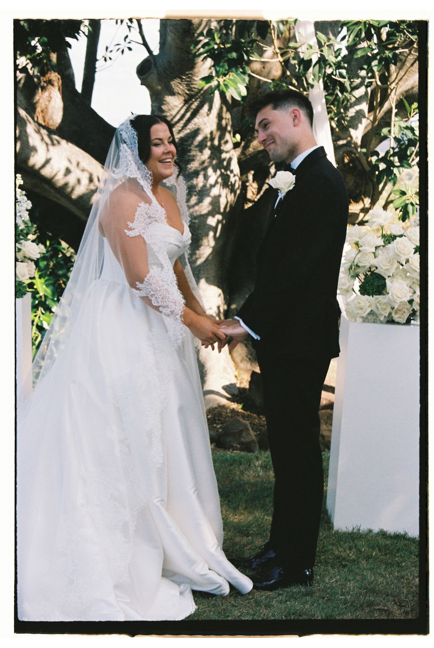 Bride and groom holding hands and smiling during outdoor wedding ceremony beside white floral arrangement.