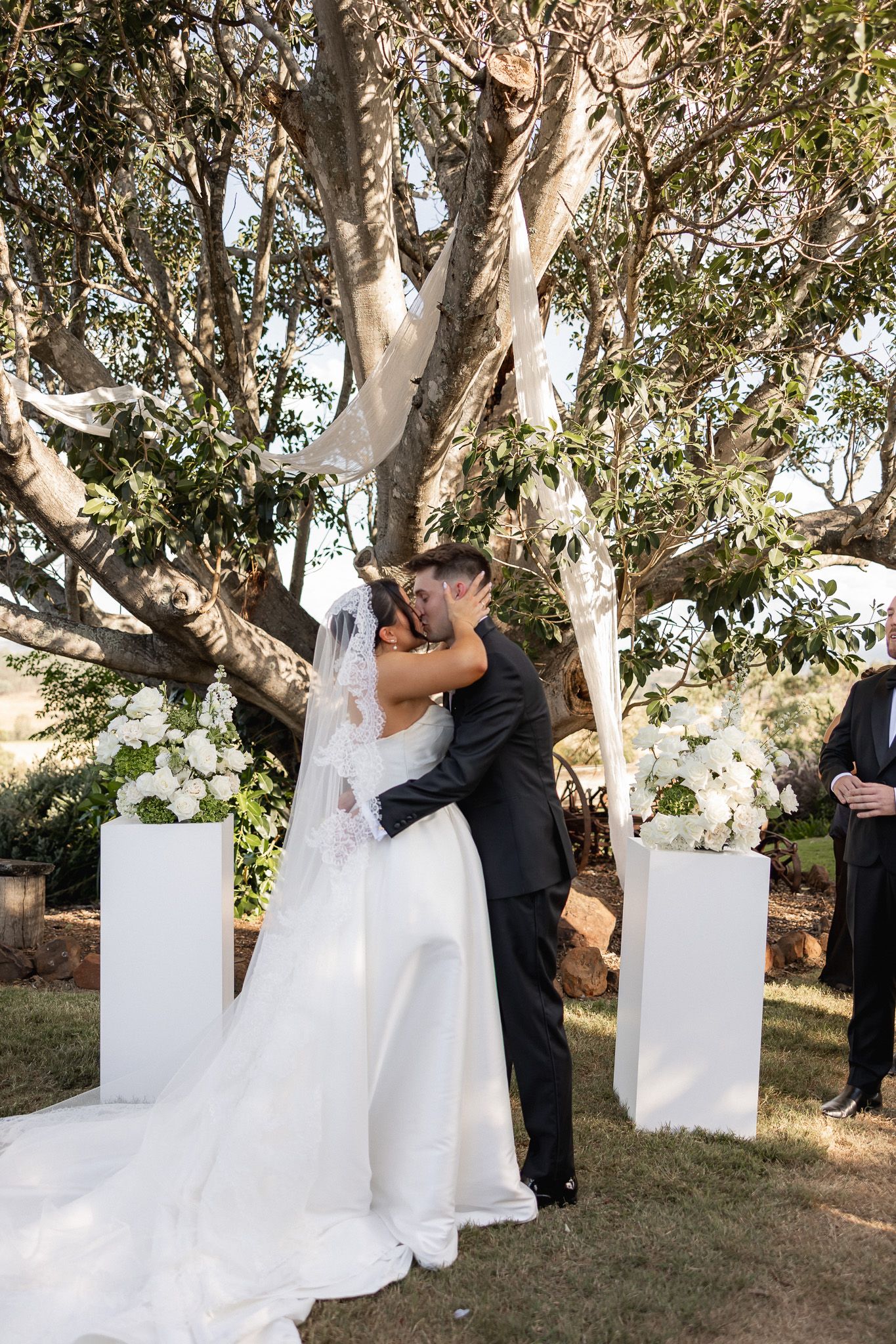 A bride and groom share their first kiss under a large tree adorned with white floral arrangements outdoors.
