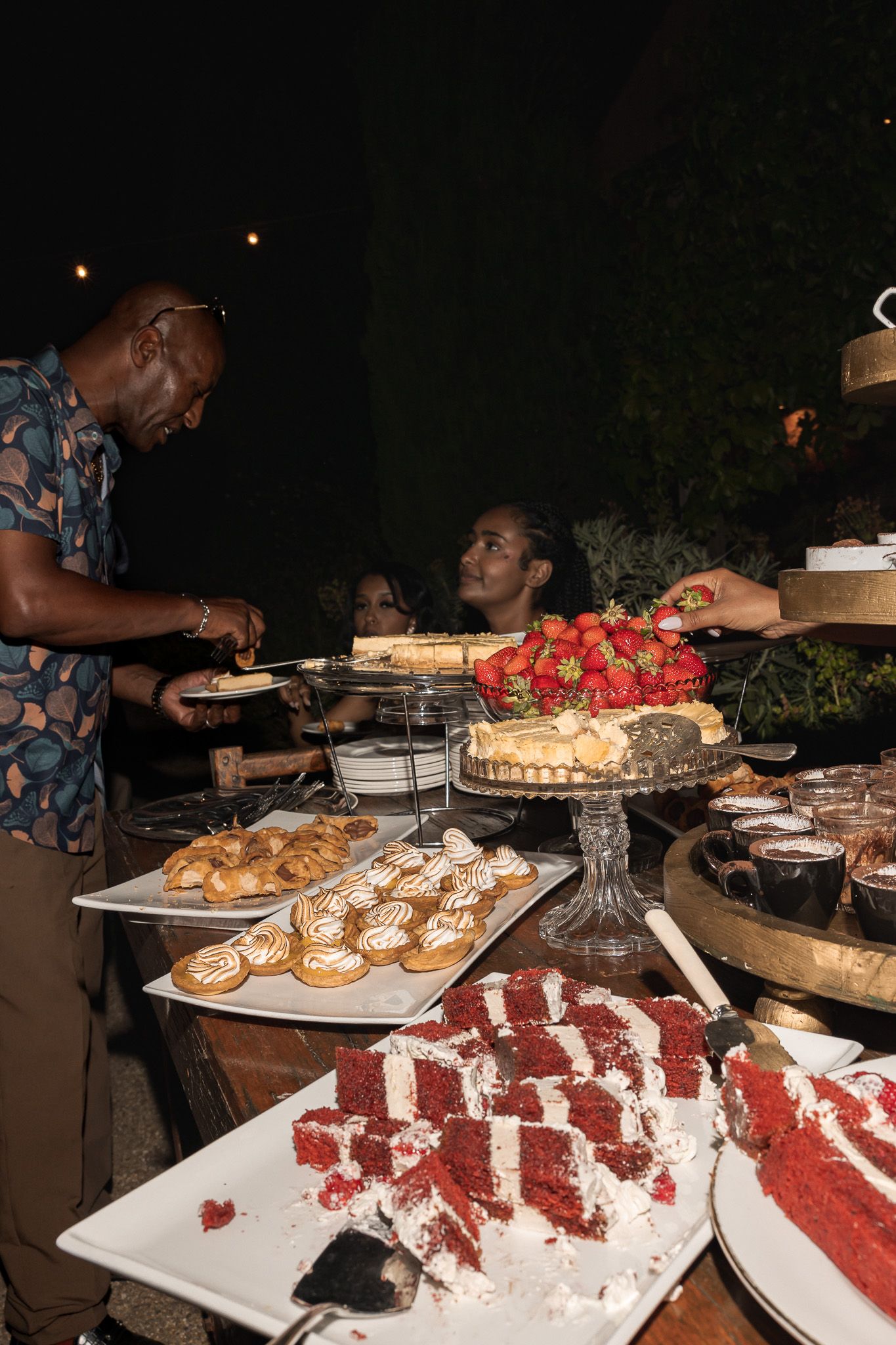 Wedding guests serving themselves from a lavish dessert table with red velvet cake and assorted sweets at night.