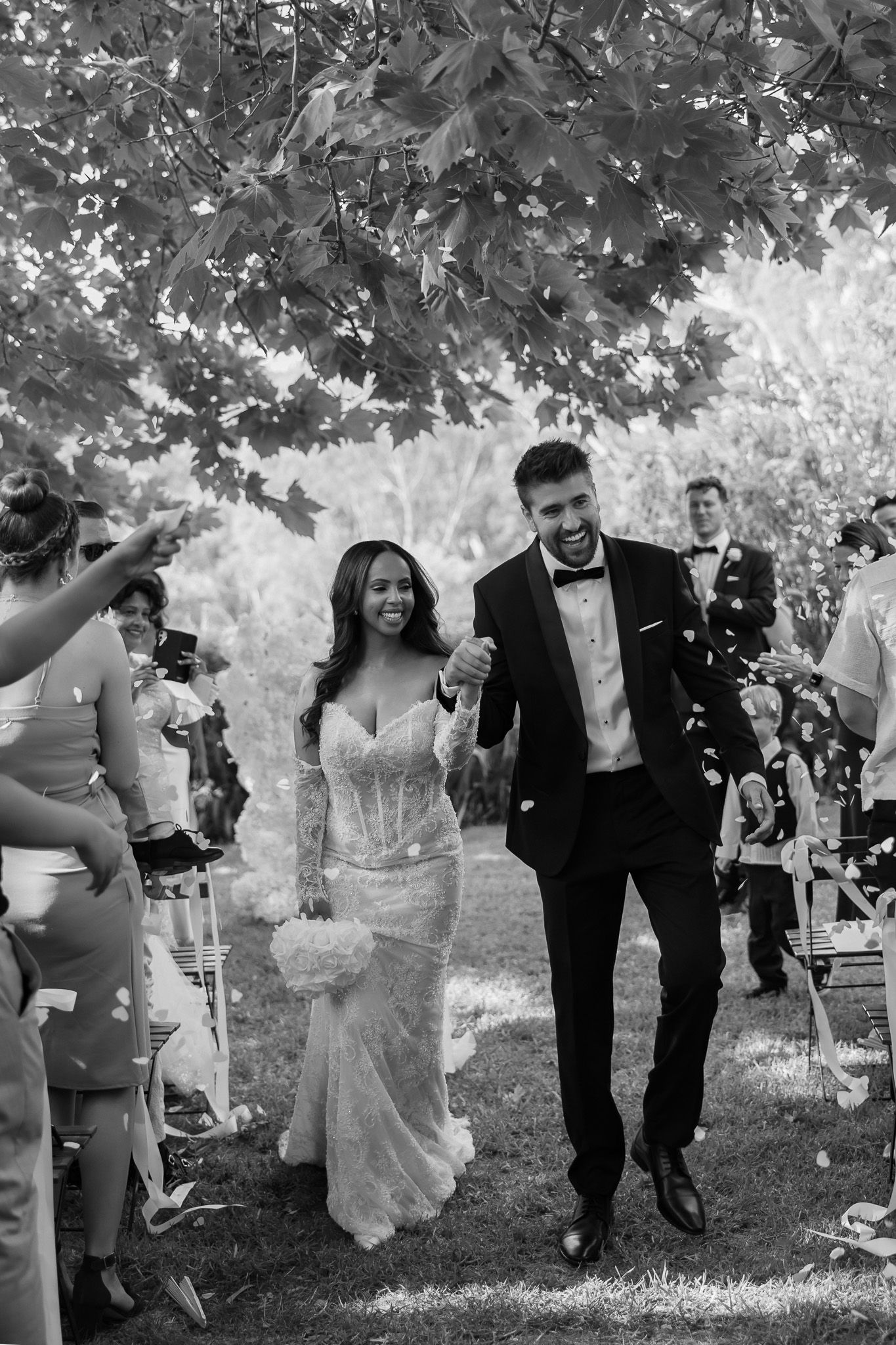 Black and white photo of joyful newlyweds walking down the aisle as guests toss confetti outdoors.