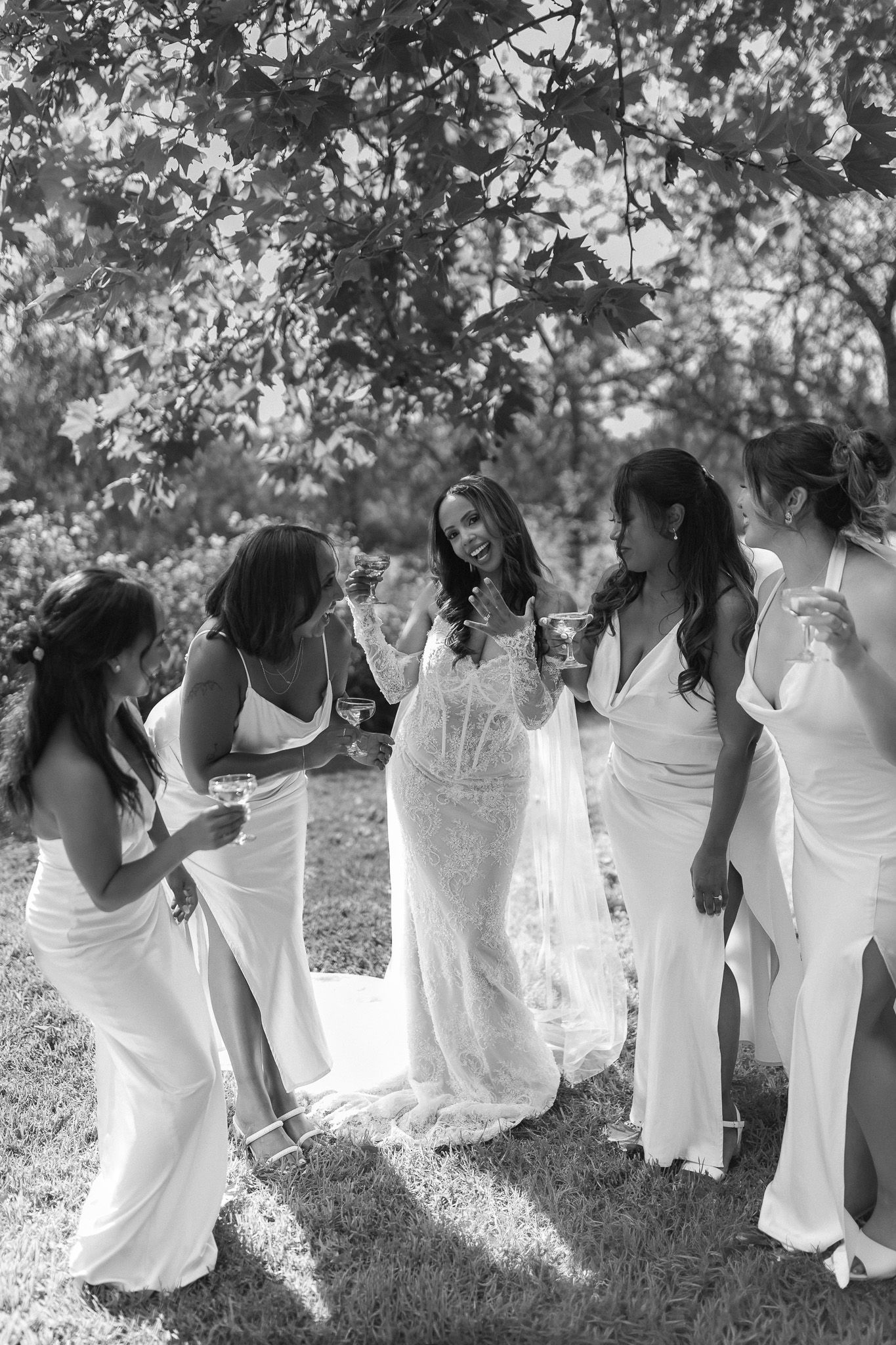 Bride in lace gown laughing with four bridesmaids in white dresses outdoors under trees, black and white photo.