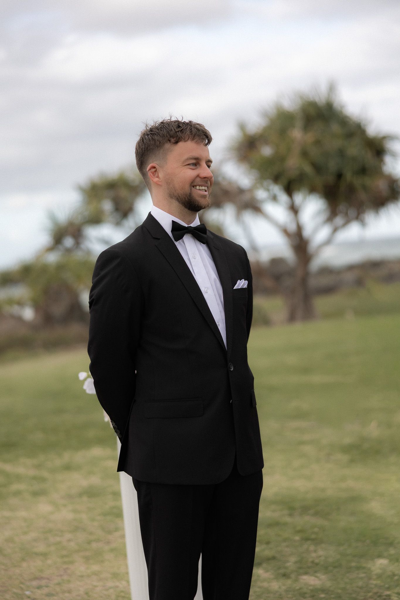 Smiling groom in black tuxedo with bow tie and white pocket square standing outdoors on green grass.