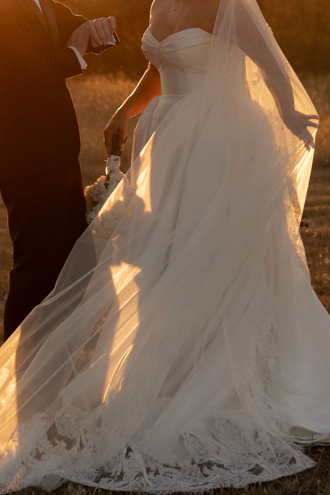Golden sunset light illuminates bride in strapless lace wedding gown holding bouquet while groom holds her veil.