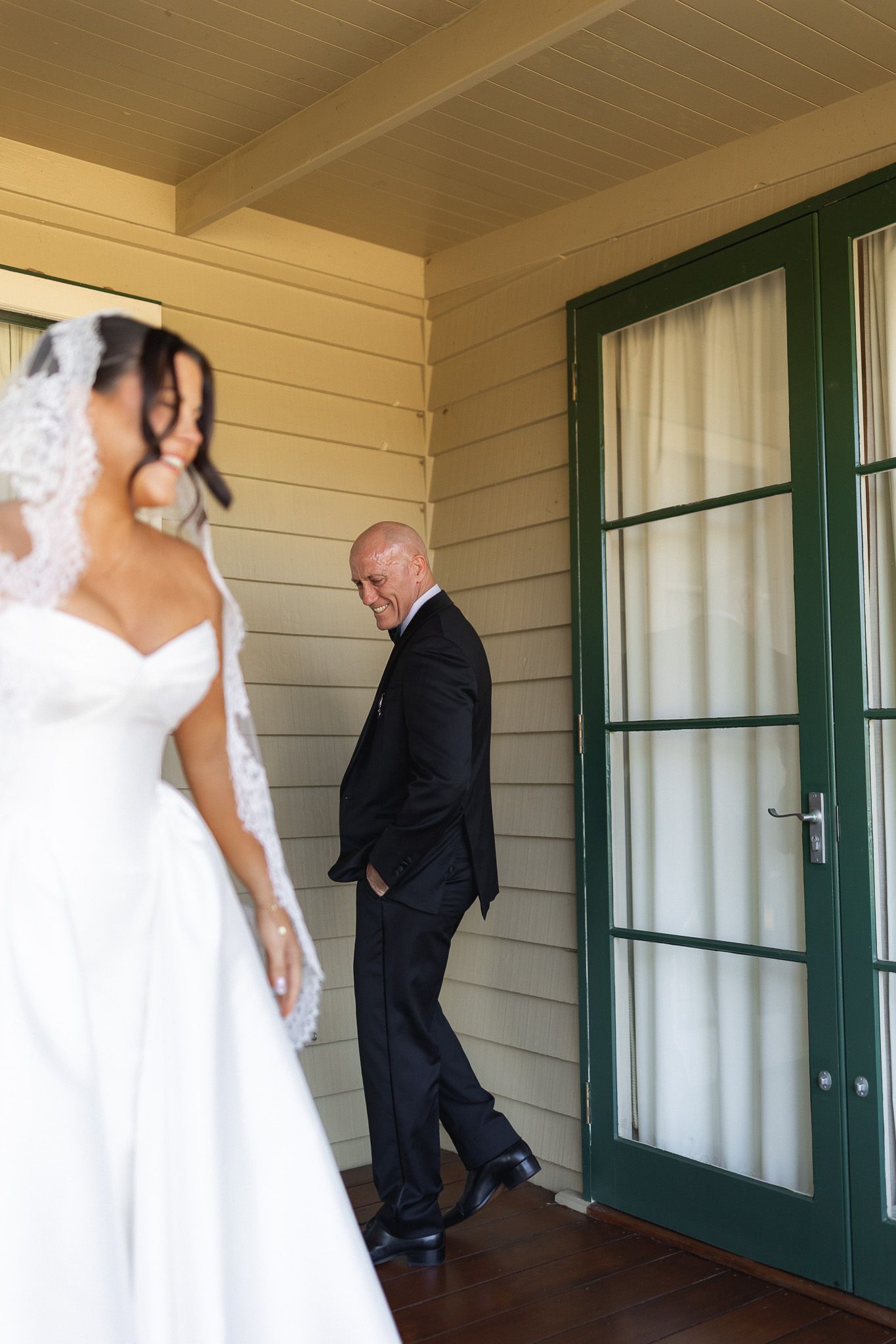 Groom in black suit watches bride in white gown and veil on porch of yellow building with green door.