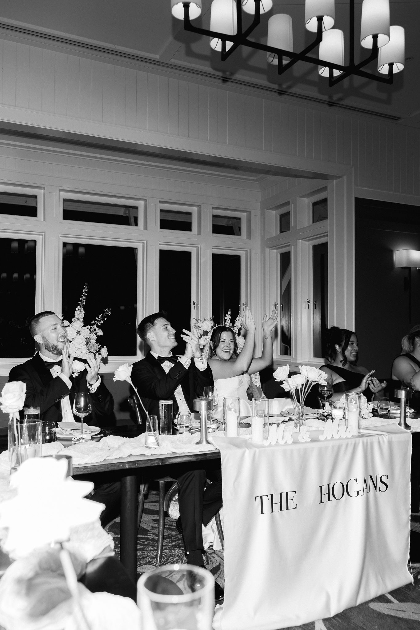 Wedding party raising glasses in a toast at the Hogans reception table in black and white.