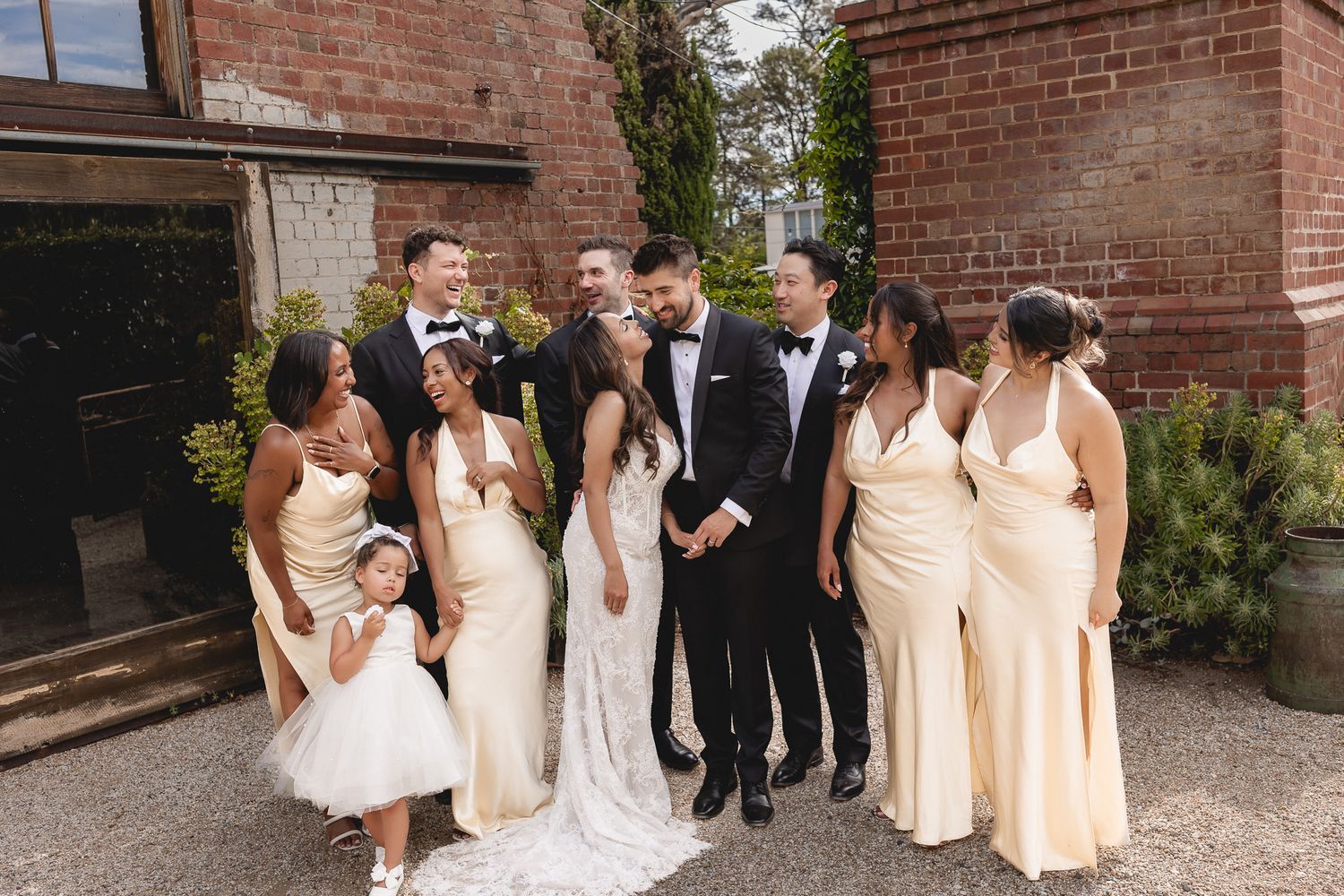 Wedding party in cream gowns and tuxedos laughing together outdoors near a brick building.