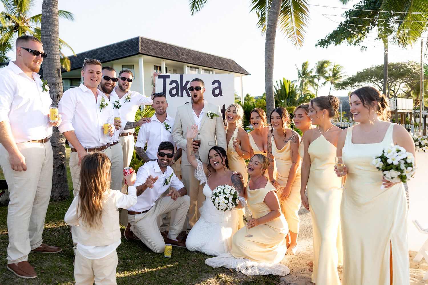 Wedding party celebrating on tropical beach with palm trees and Takoa sign in background.