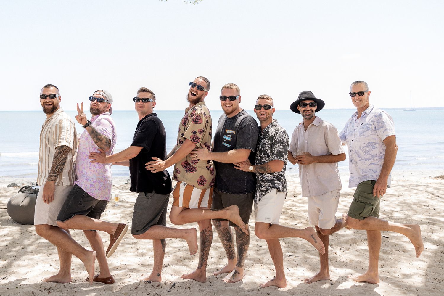Group of eight men posing on a sunny beach, wearing casual summer outfits, smiling and lifting their legs.