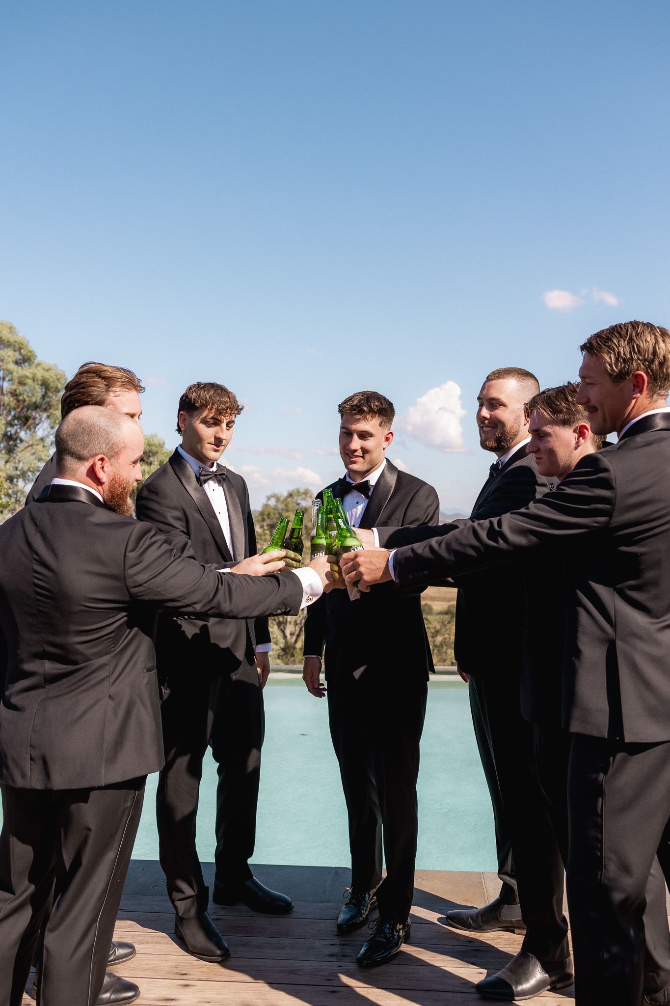 Groomsmen in black tuxedos toasting with beer bottles outdoors under a clear blue sky.