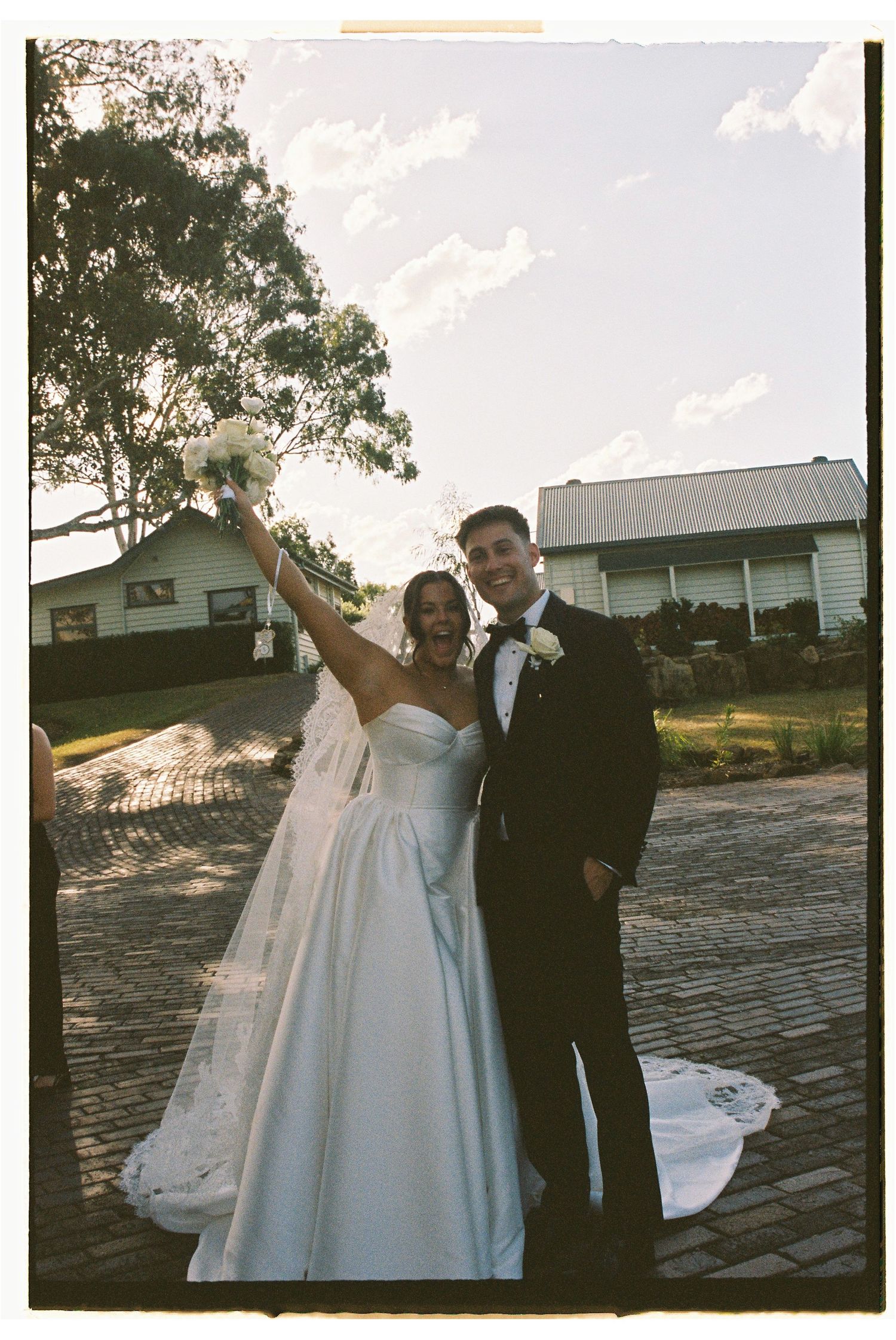 Joyful bride raises bouquet while groom embraces her on brick path outside wedding venue.