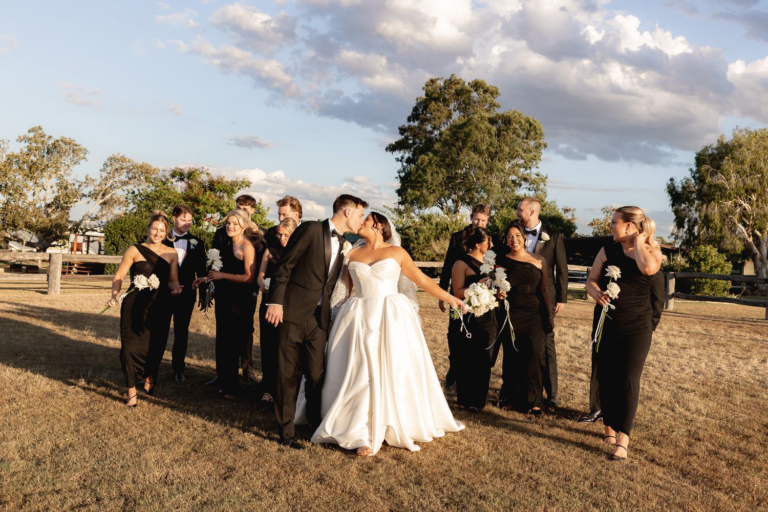 Bride and groom kissing surrounded by wedding party in black attire on a golden field.