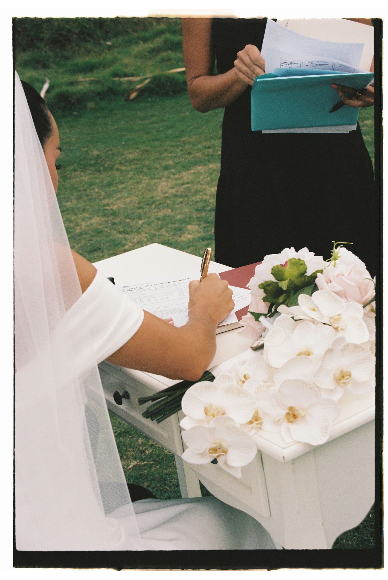 Bride in white veil signing wedding documents at a flower-adorned table during an outdoor ceremony.