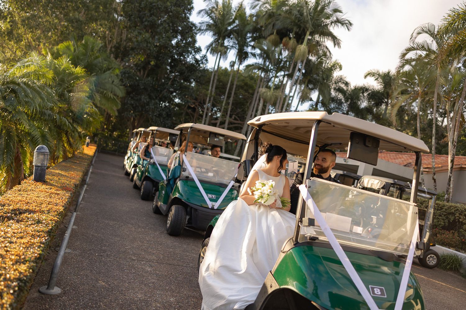 Bride in white gown boarding a green golf cart at a tropical wedding venue lined with palm trees.