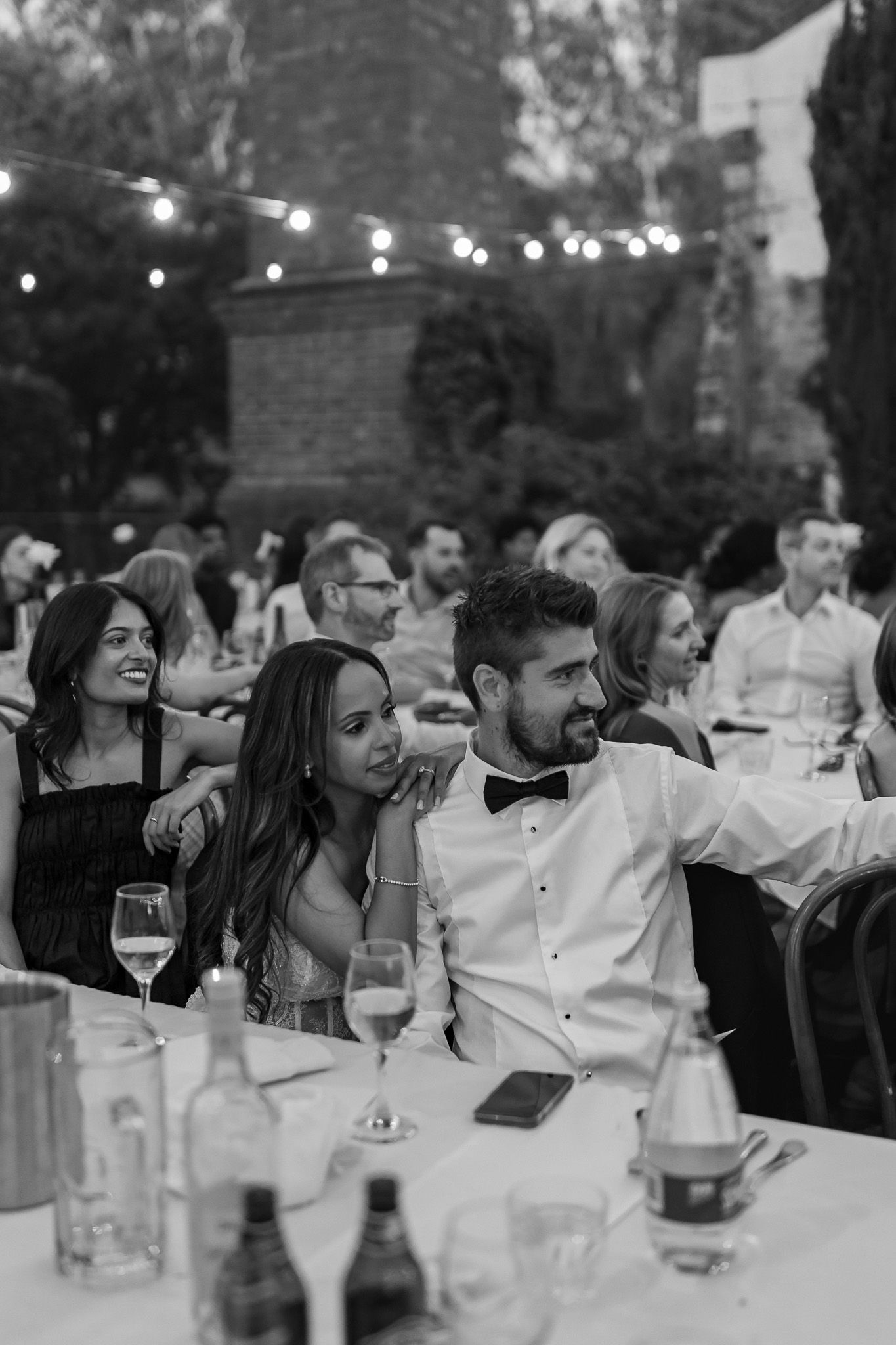 Black and white photo of wedding guests seated at outdoor reception table under string lights at dusk.