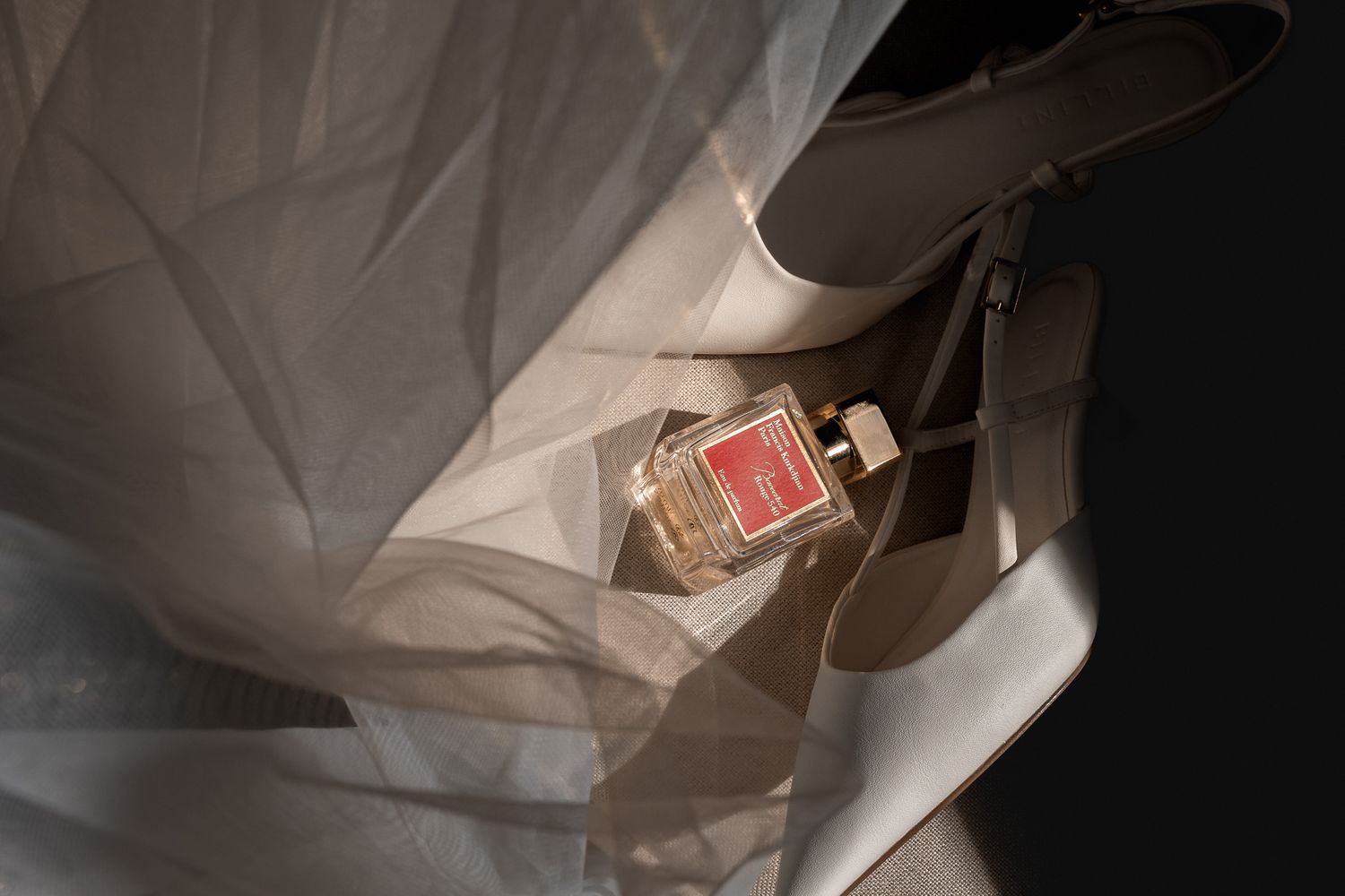 White bridal heels and perfume bottle on flowing wedding veil fabric in soft natural light.