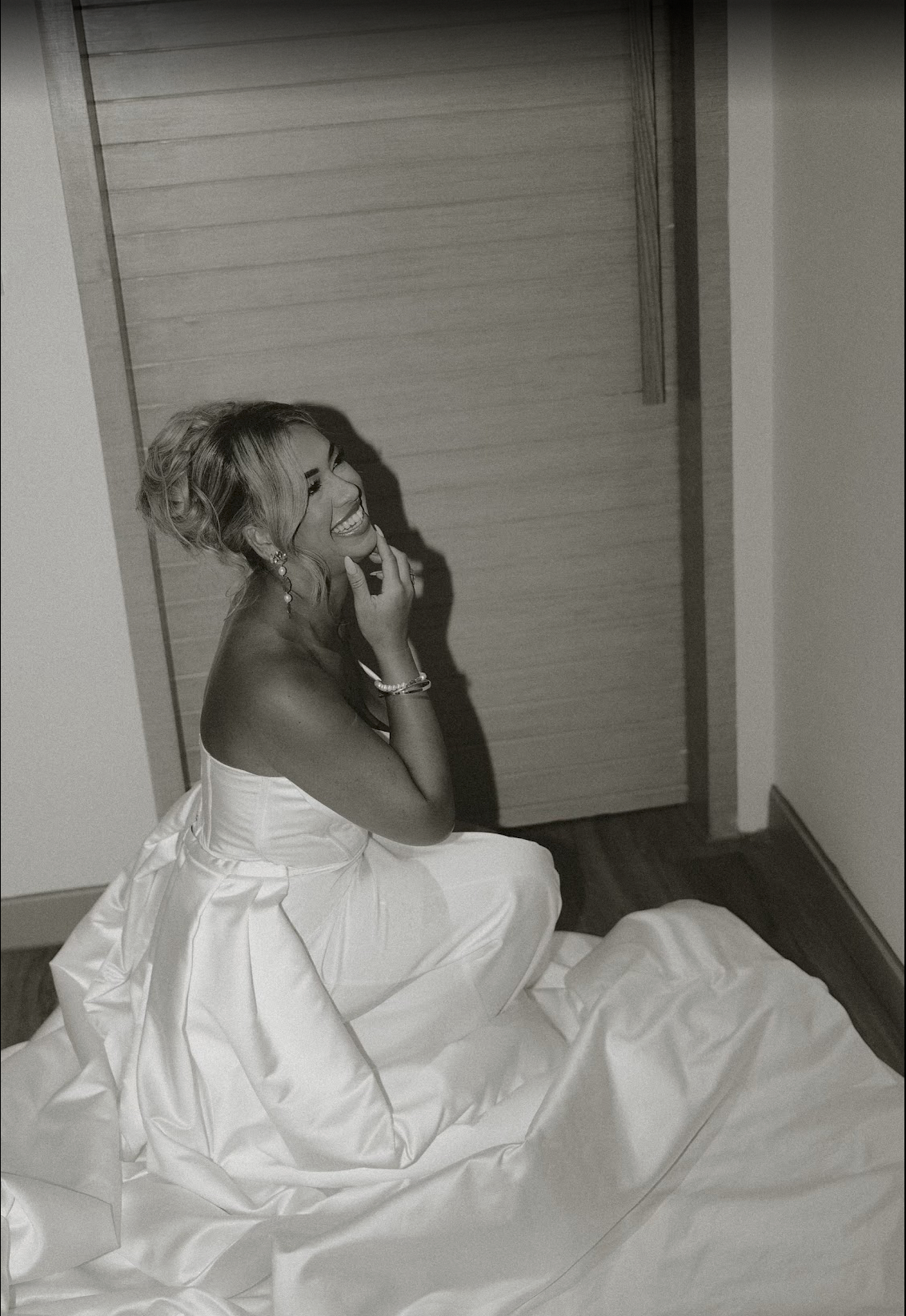 A bride in a white strapless gown sits on the floor laughing, captured in elegant black and white photography.