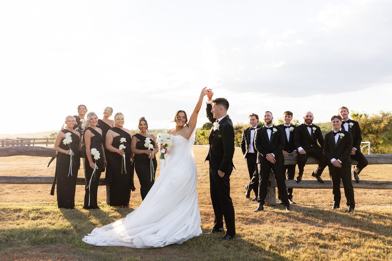 Wedding party celebrates outdoors, bride and groom raise hands while bridesmaids in black dresses and groomsmen in tuxedos cheer.