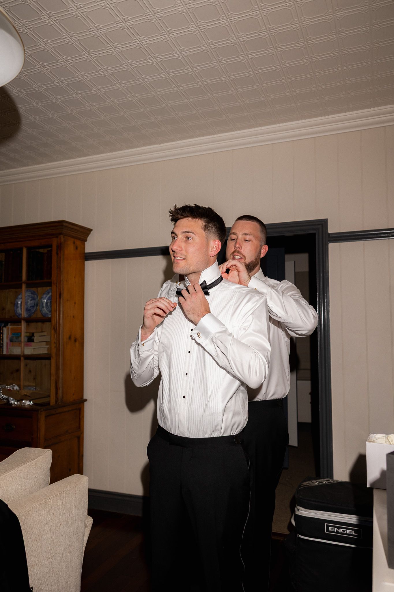 Groom in white dress shirt having bow tie adjusted by groomsman while getting ready indoors.