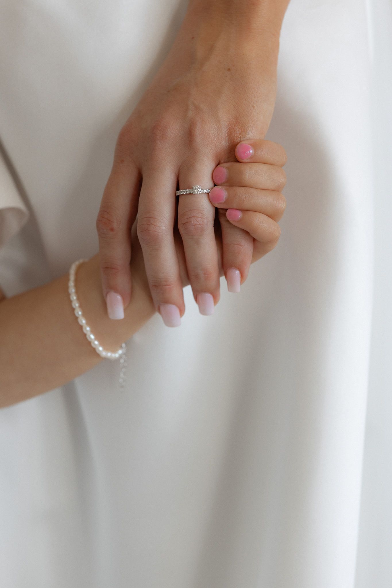 Close-up of bride's hands showing diamond engagement ring and pearl bracelet against white wedding dress.