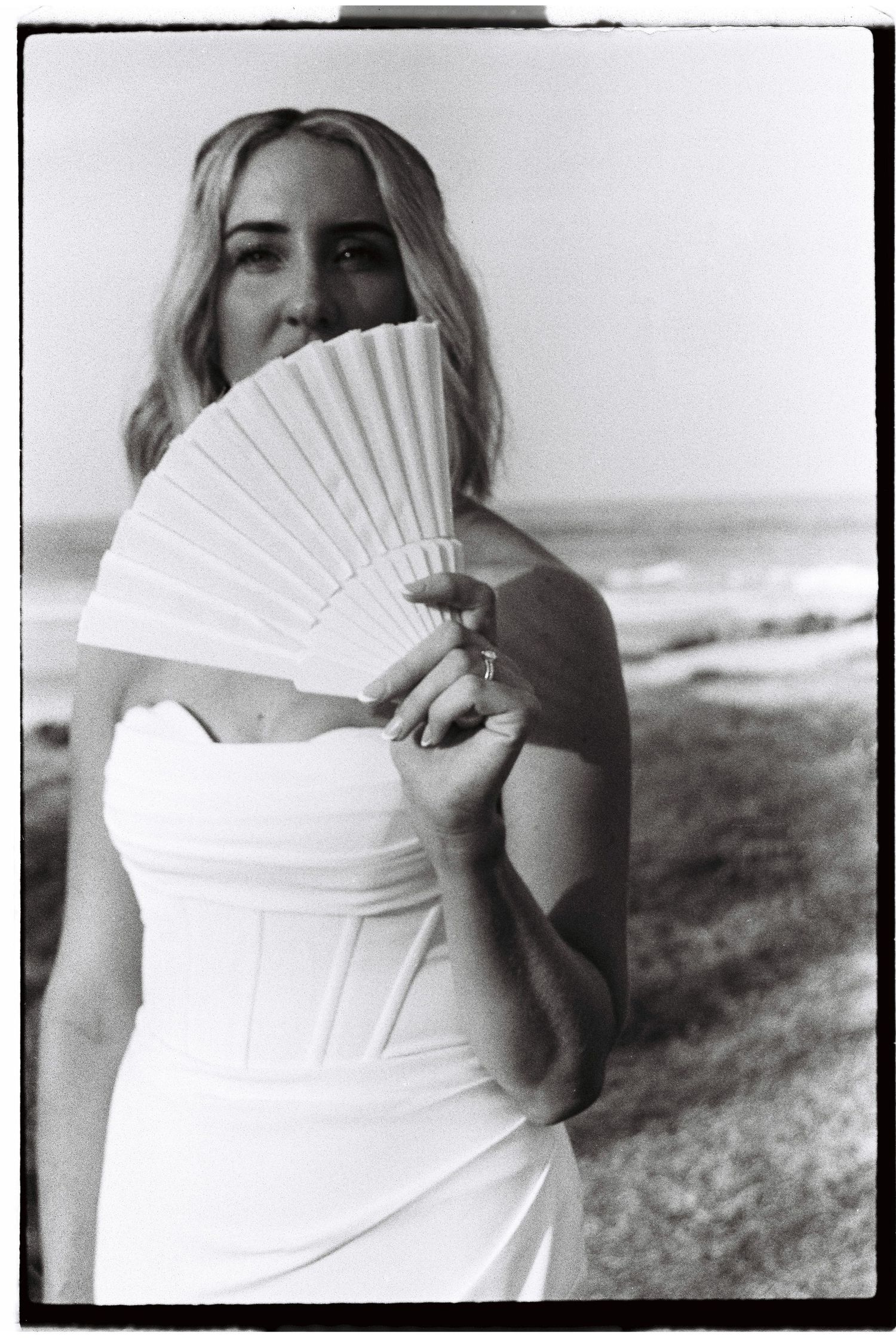 Bride in white strapless gown holding decorative fan near beach in black and white photo.