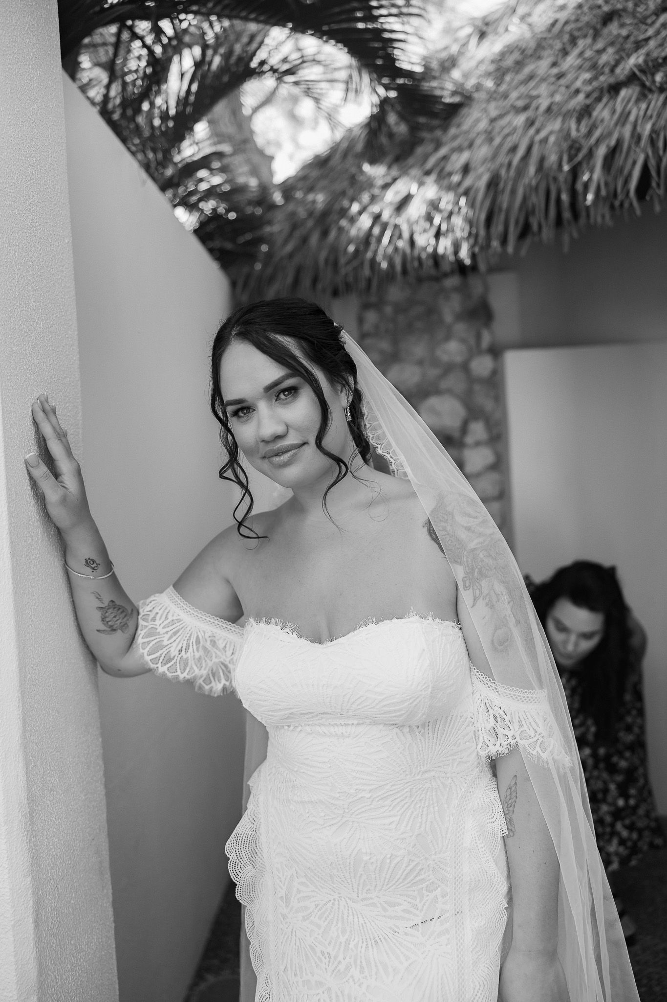 Black and white photo of a bride in a lace off-shoulder gown with veil, posing gracefully against a wall outdoors.