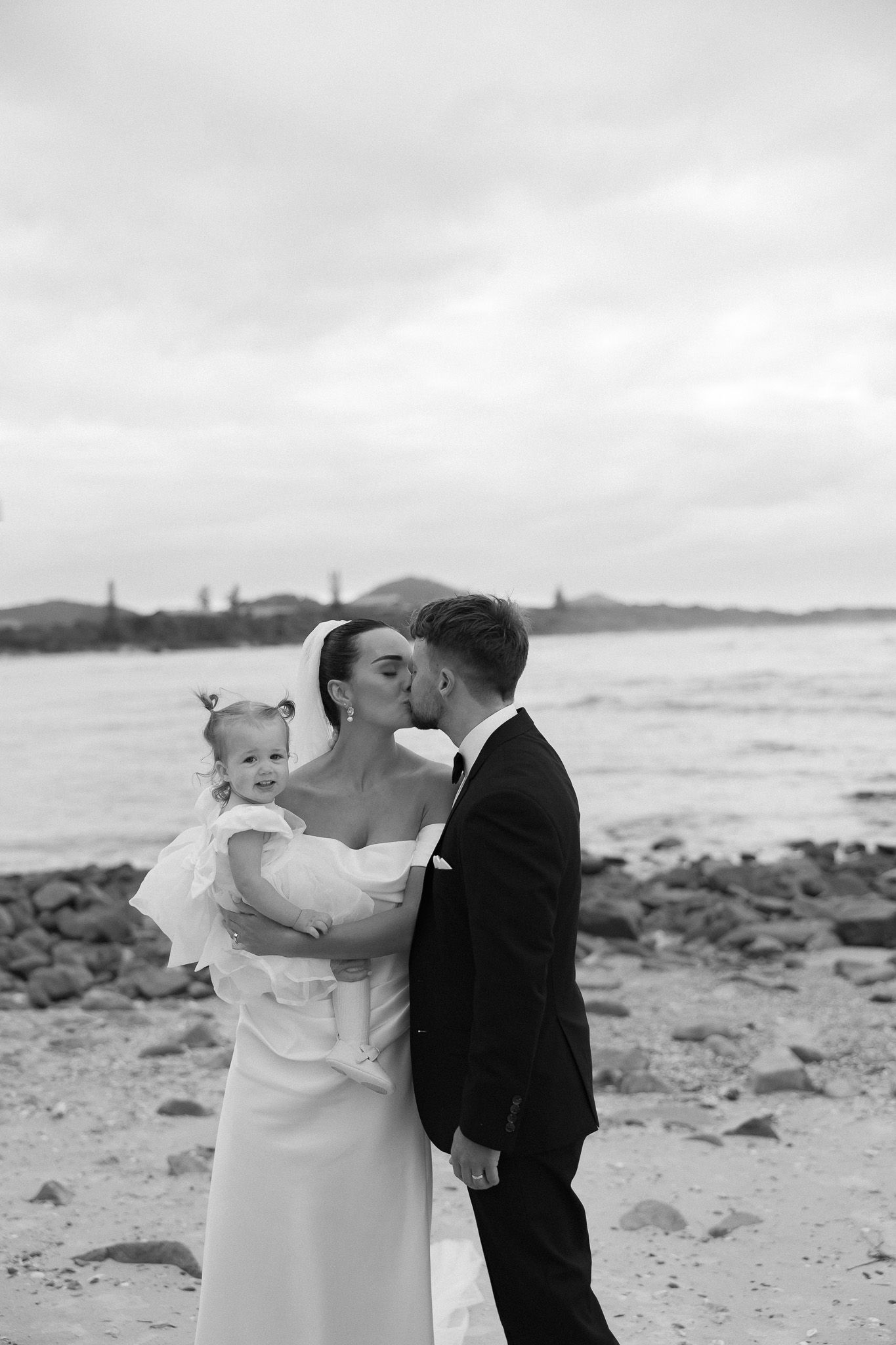 Black and white photo of bride and groom kissing on beach while holding their baby in wedding attire.