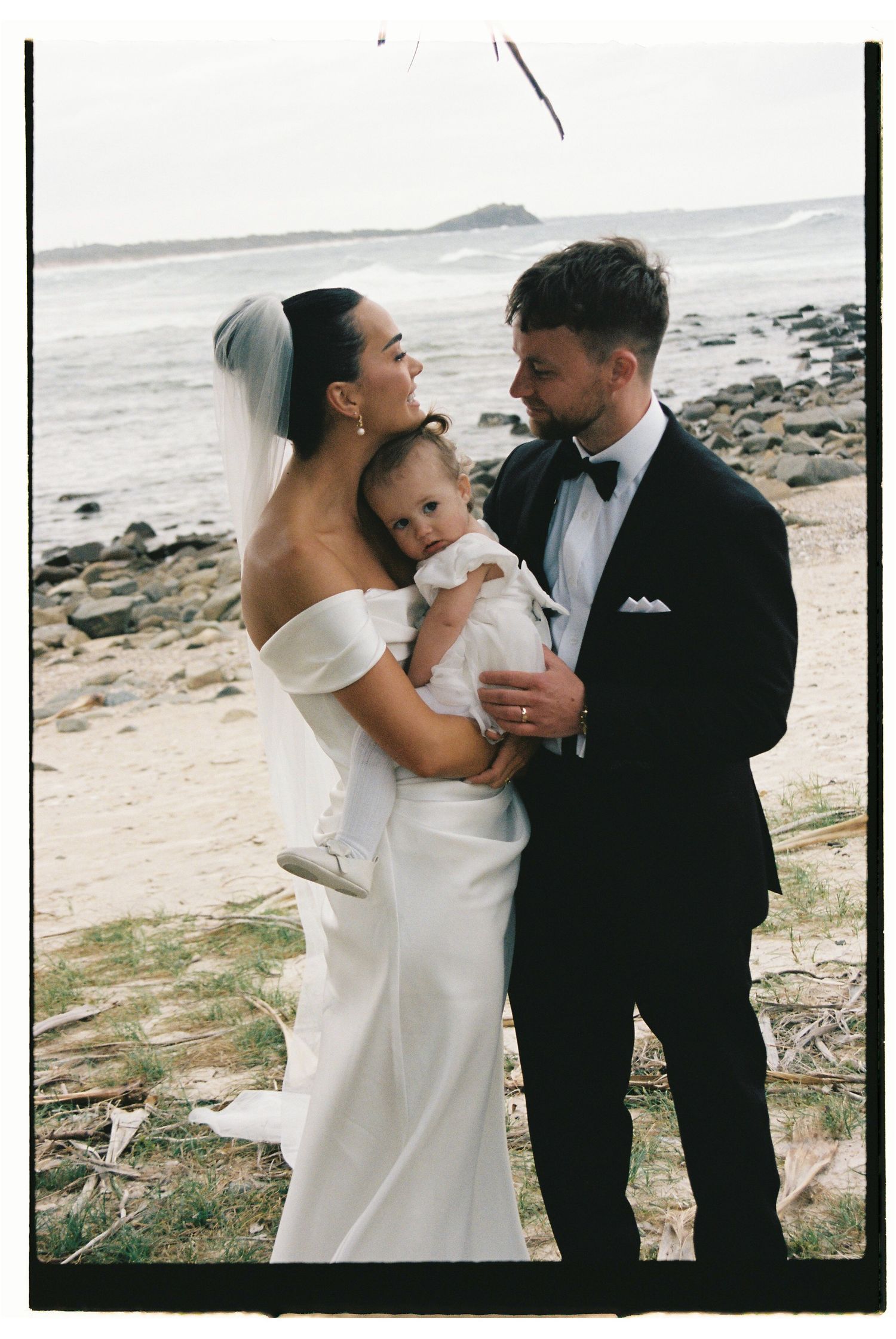 Bride and groom holding baby on rocky beach, bride in off-shoulder gown with veil.