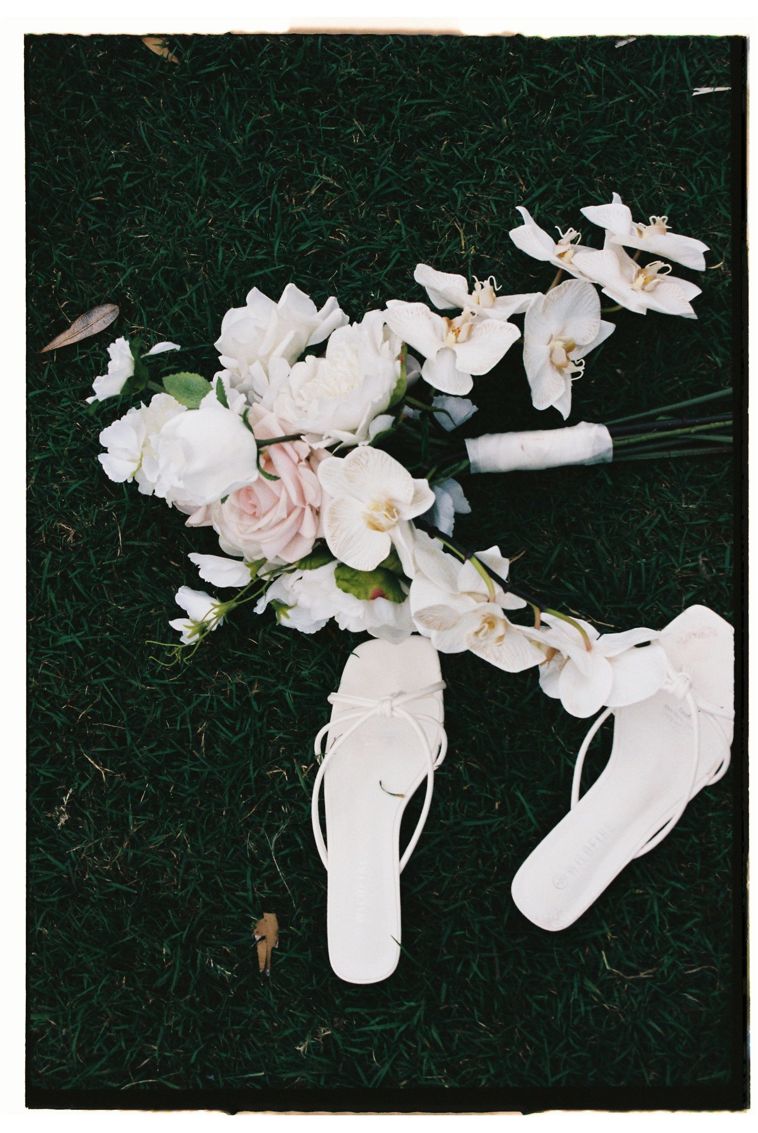 White bridal bouquet of roses and orchids beside white flip flop sandals on green grass.