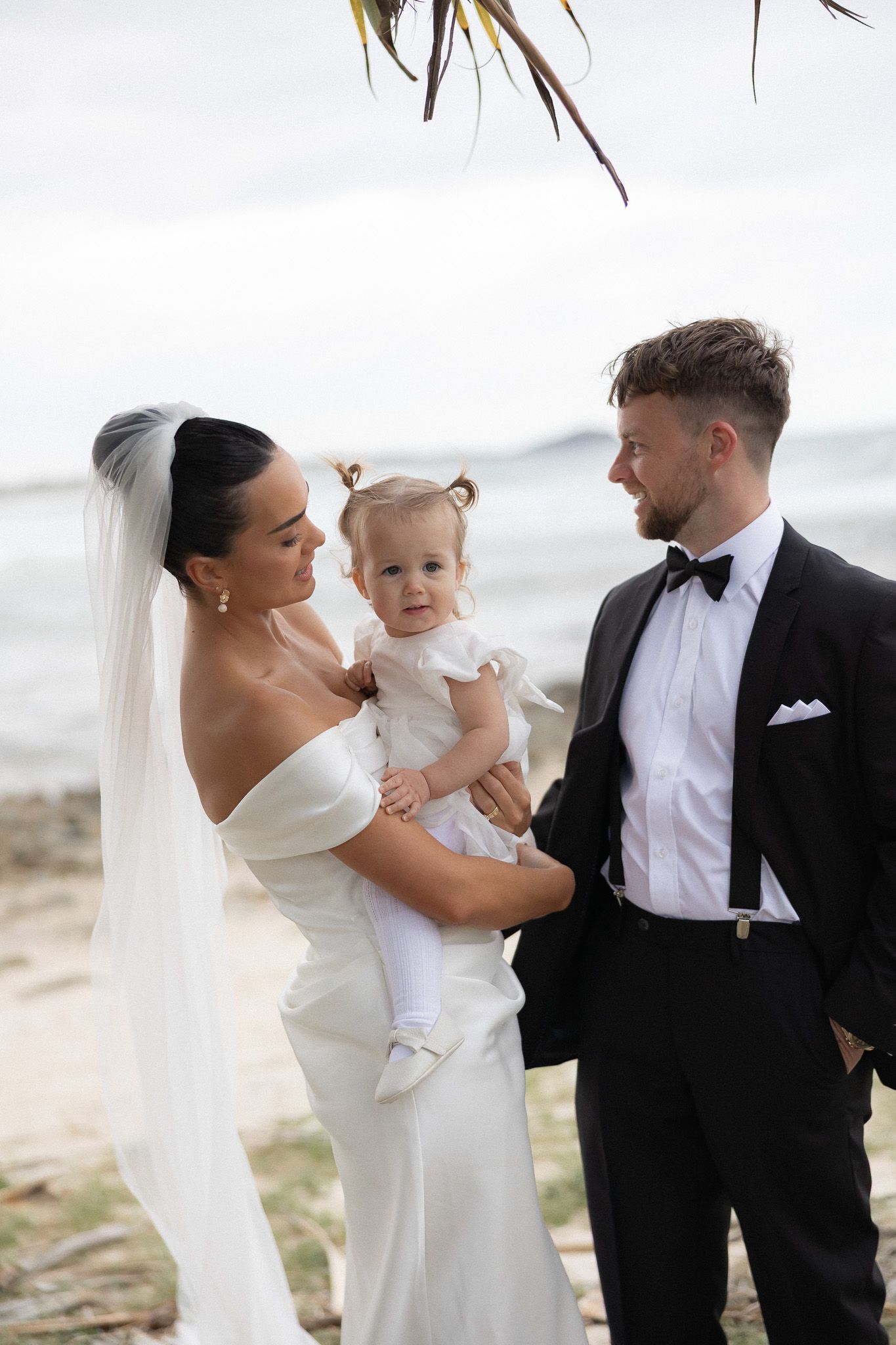 Bride in white off-shoulder gown and veil with groom and baby at beachside wedding.