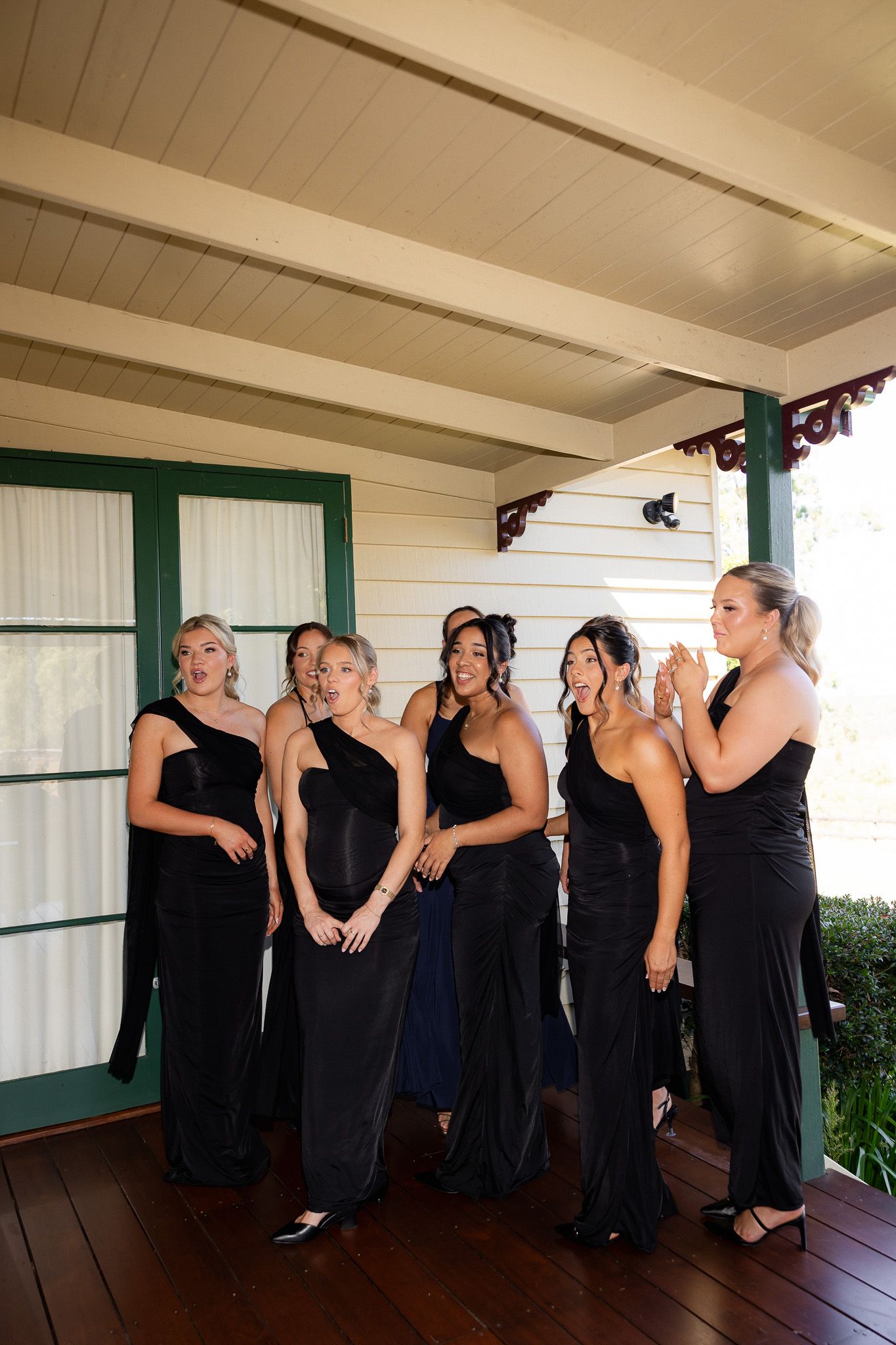 Six bridesmaids in black gowns react with excitement on a wooden porch of a white heritage building.