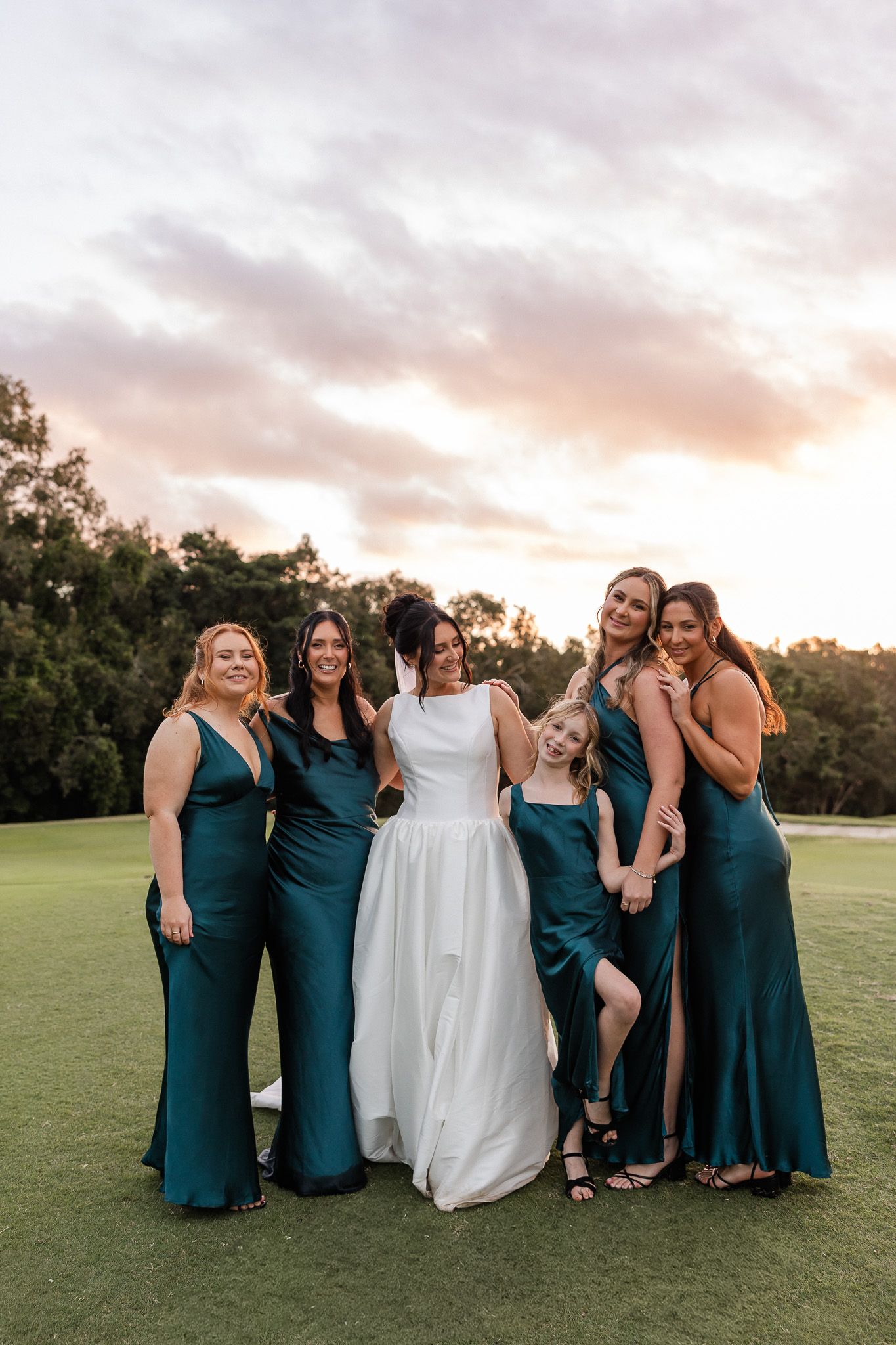 Bride in white gown poses with five bridesmaids in emerald green satin dresses on a golf course at sunset.