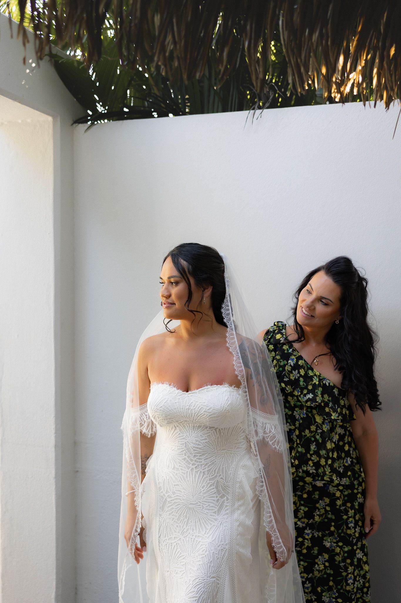 Bride in lace wedding gown and veil stands with bridesmaid in floral dress against white wall.