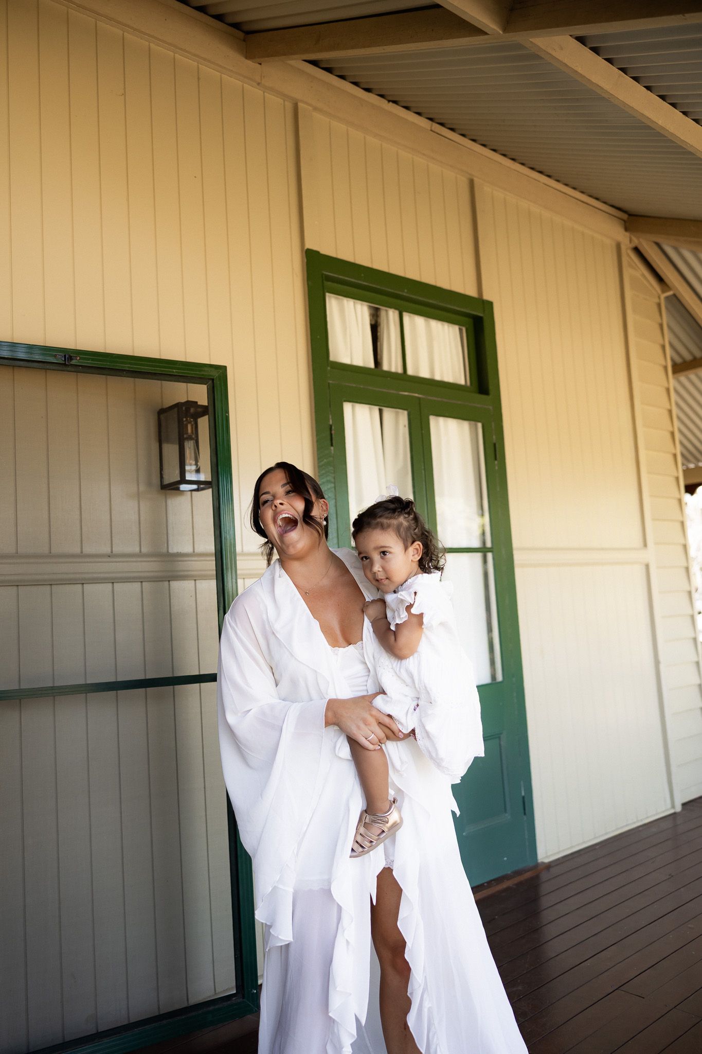 A bride in a white robe holds a young child in white on a wooden porch with green-trimmed doors.