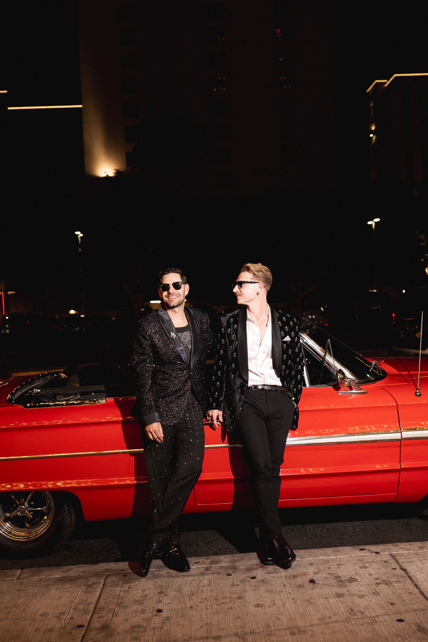 Two grooms in sparkly black tuxedo jackets leaning against a classic red convertible at night.