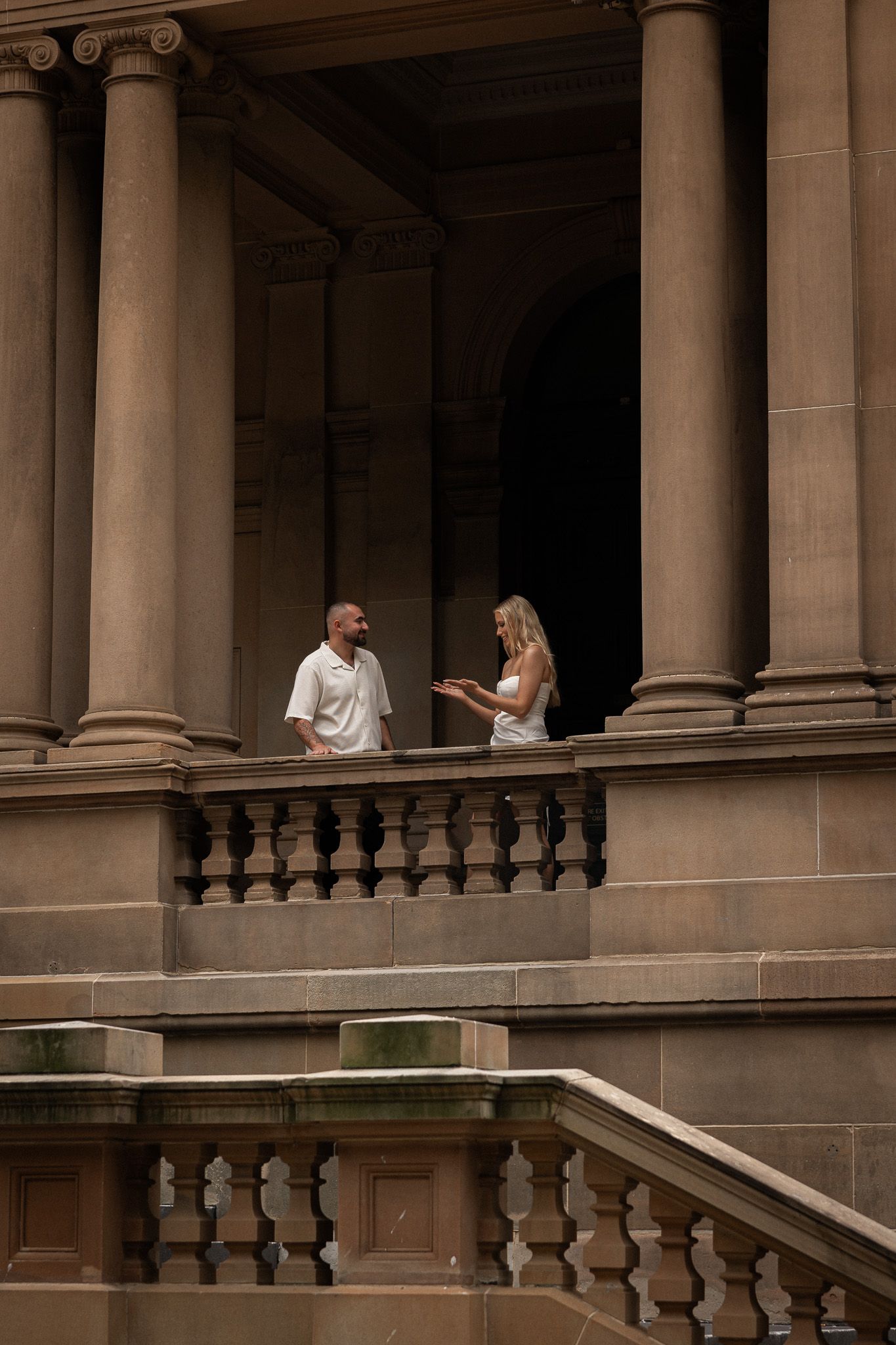 Couple in white attire standing on ornate stone balcony with classical columns of historic building.