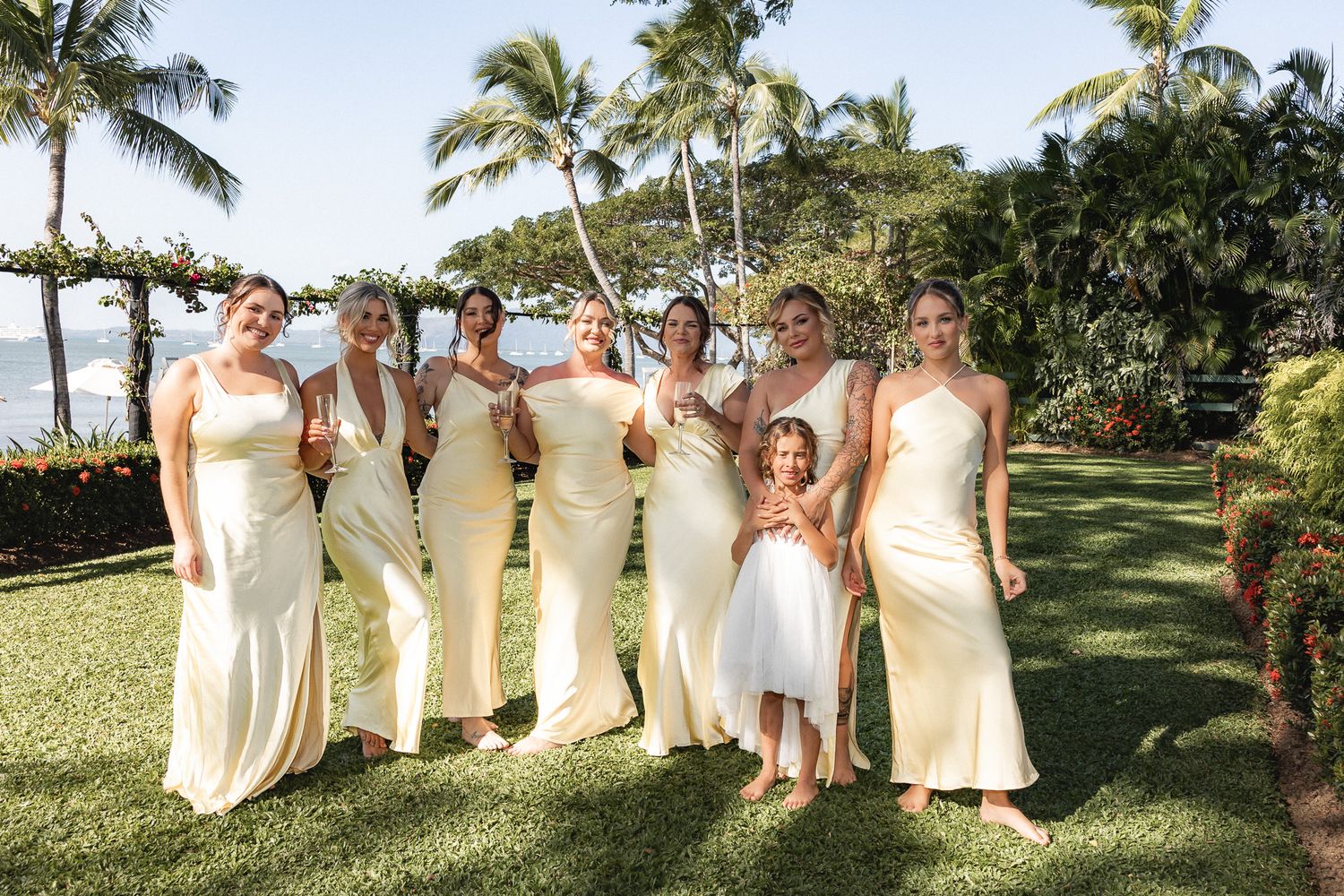 Bridesmaids in ivory gowns pose with flower girl on tropical lawn with palm trees and ocean backdrop.