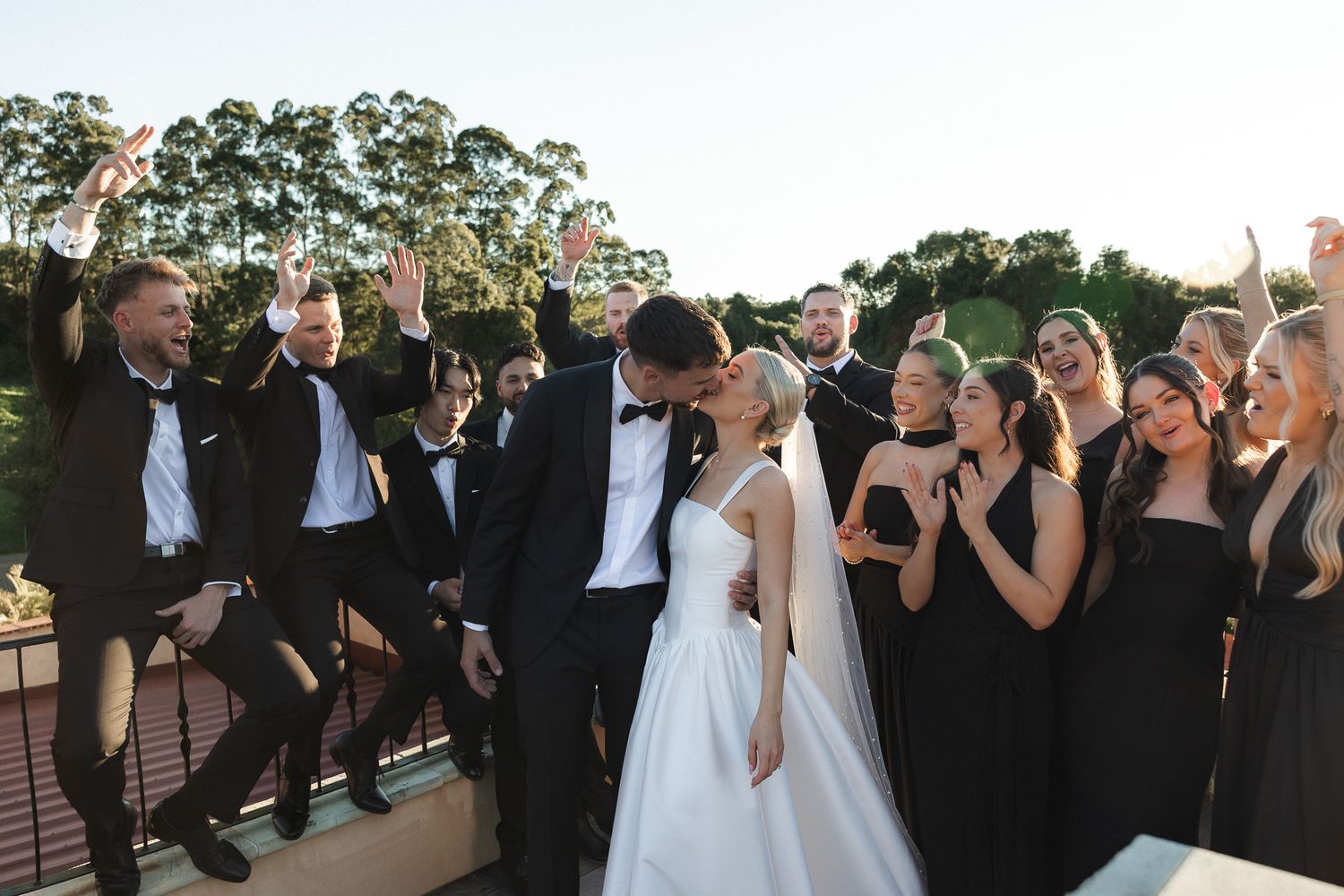 Bride and groom kissing surrounded by cheering wedding party in black attire at outdoor wedding celebration.