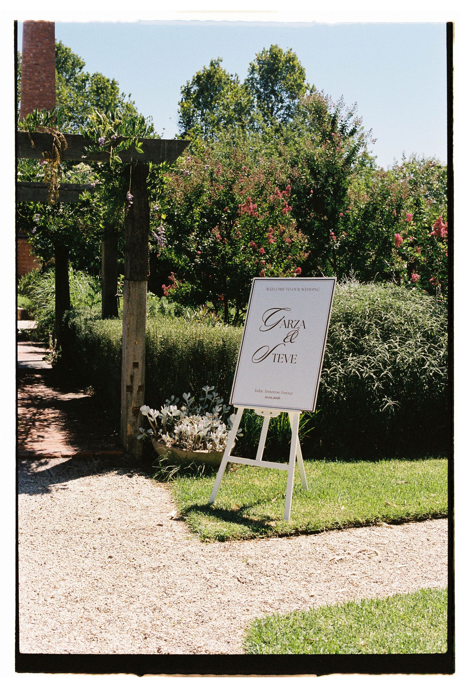 Elegant white easel wedding welcome sign for Garza and Steve in a lush garden setting on a sunny day.