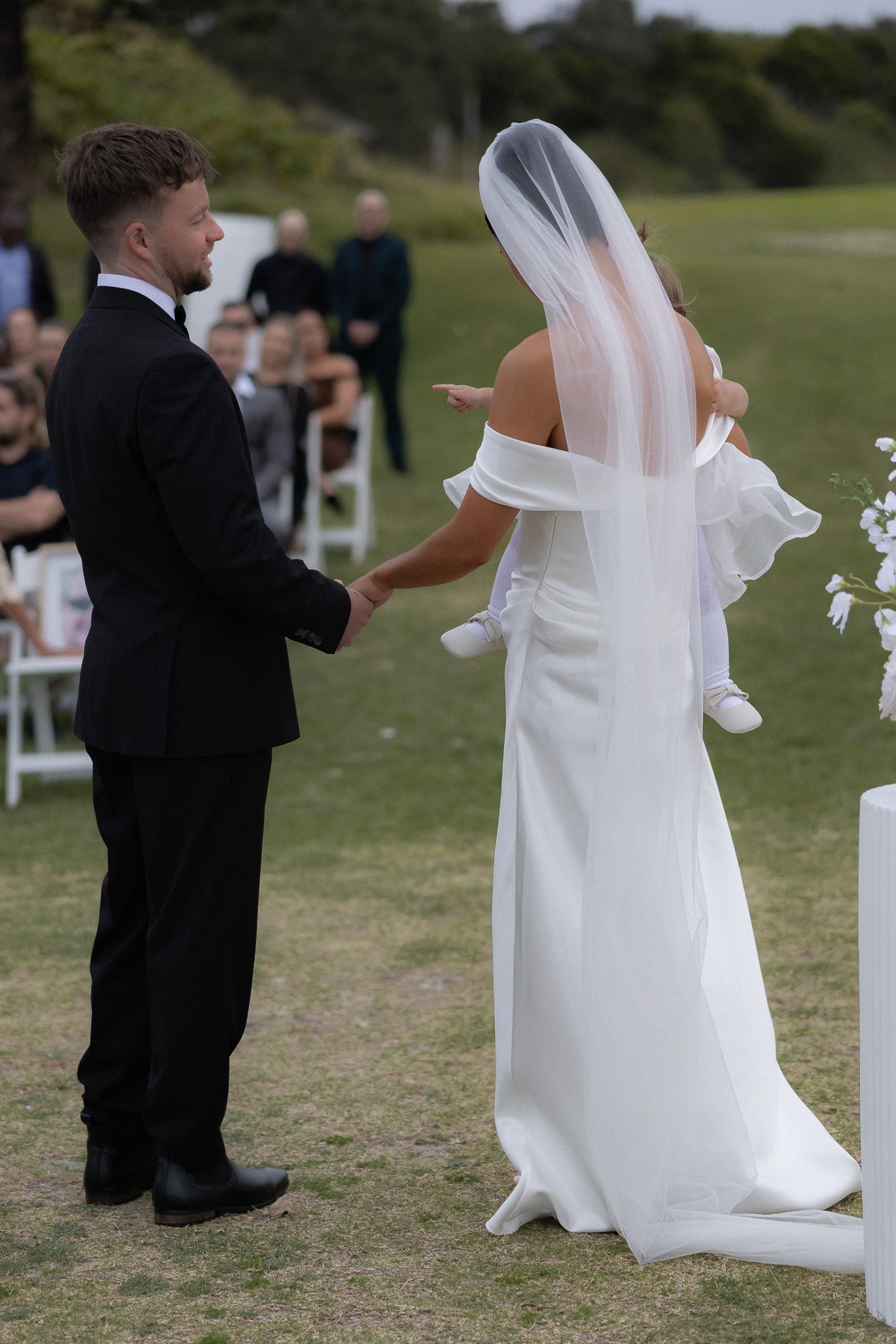 Bride in white off-shoulder gown and veil holds hands with groom in black suit during outdoor wedding ceremony.