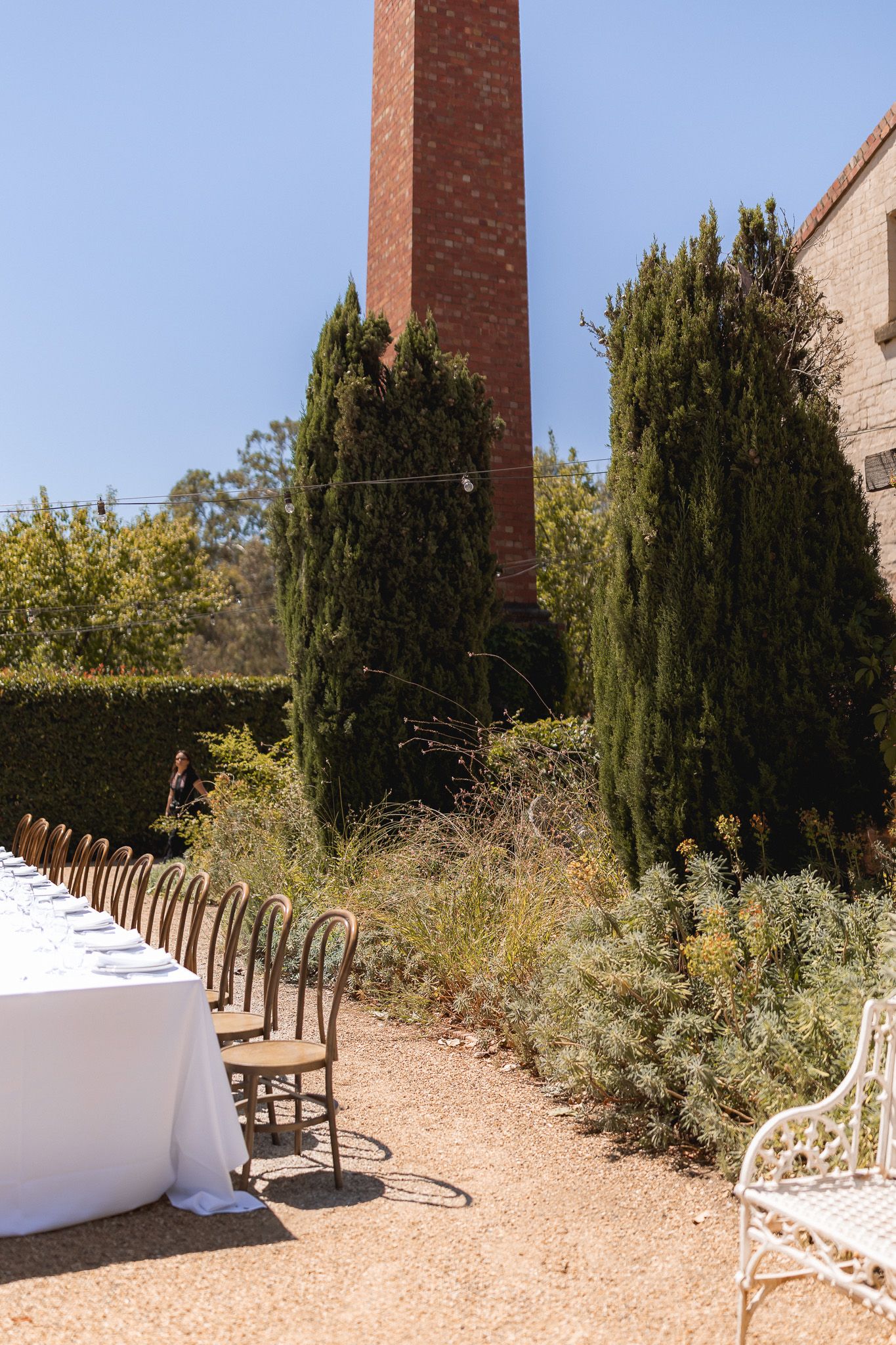 Elegant outdoor wedding reception with long white-draped table, bentwood chairs, cypress trees, and a brick chimney under blue sky.