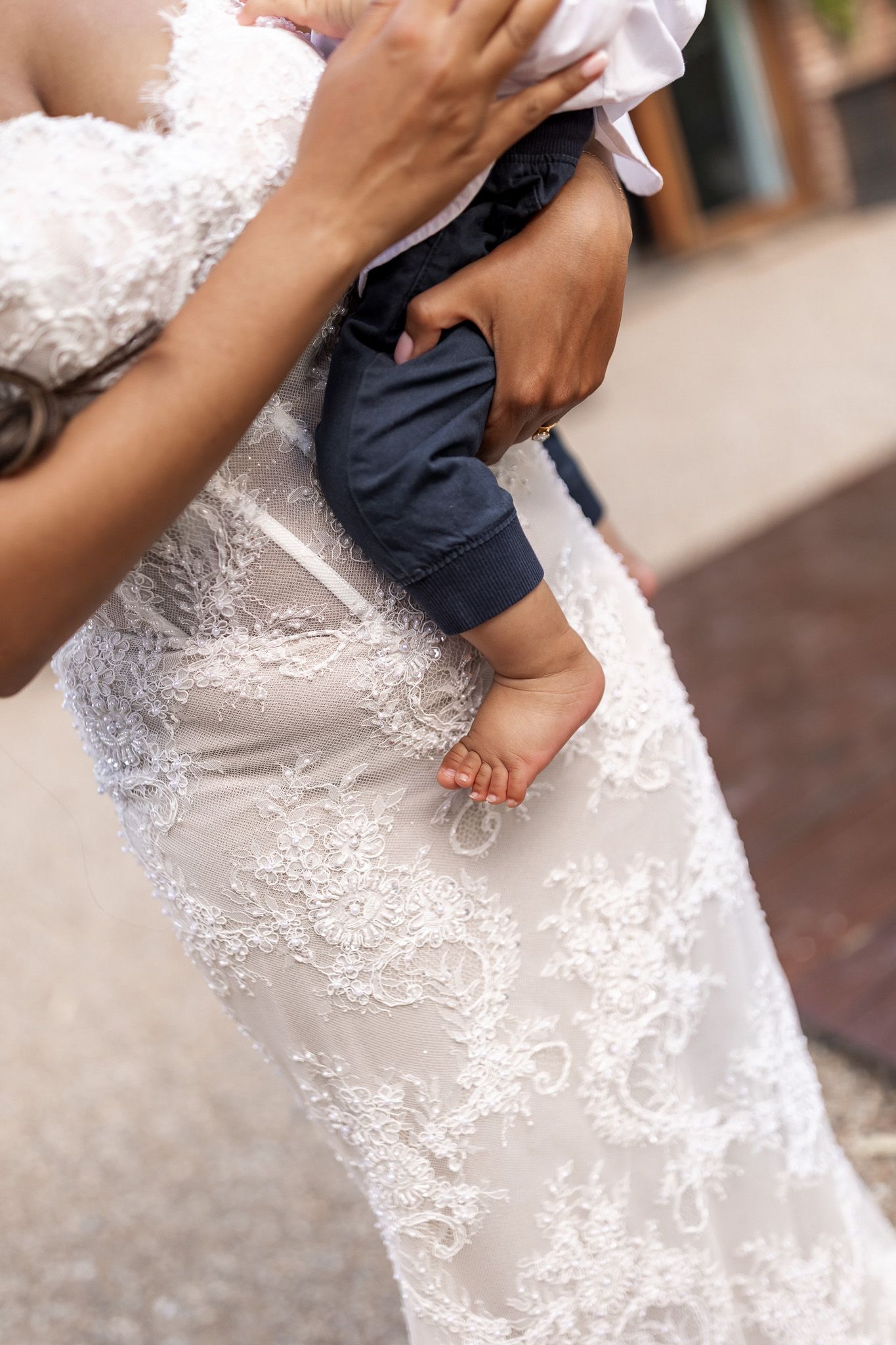 Close-up of bride in intricate lace wedding dress holding a baby with tiny bare feet visible.
