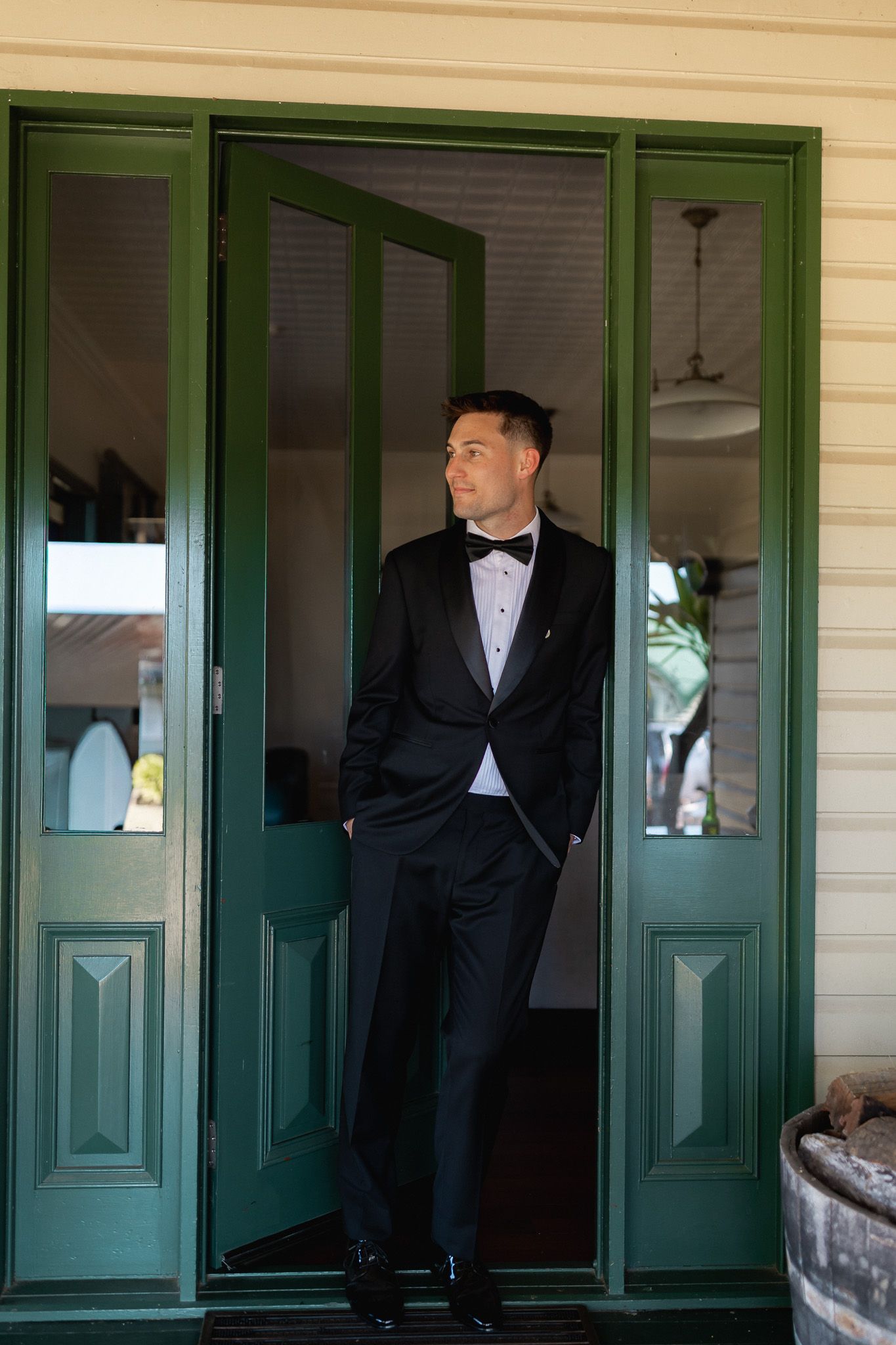 Elegant groom in black tuxedo and bow tie standing in green double doors on wedding day.