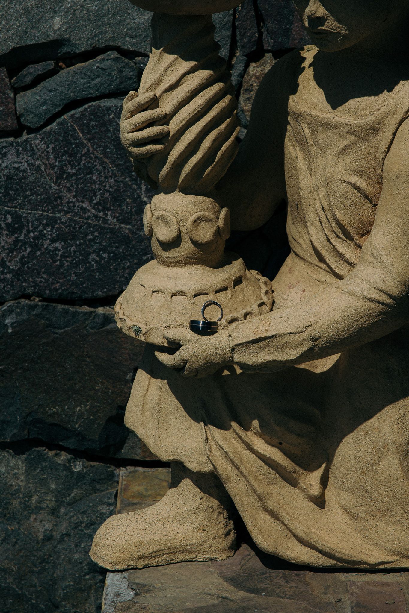 Stone statue holding wedding ring on ornate carved pedestal against dark rock wall.