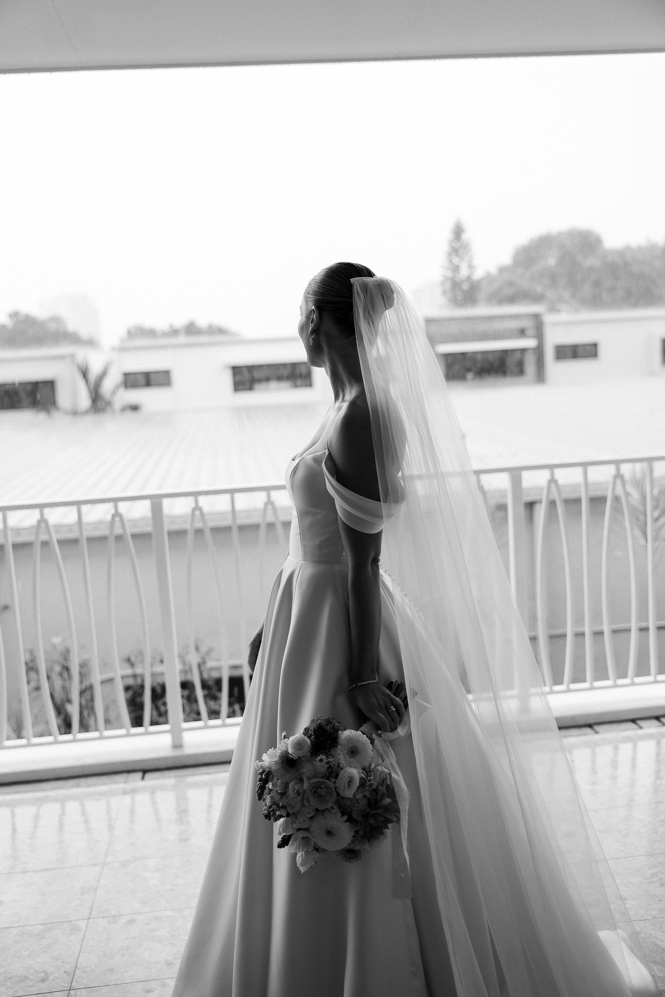Black and white photo of bride in ballgown and veil holding bouquet on balcony.