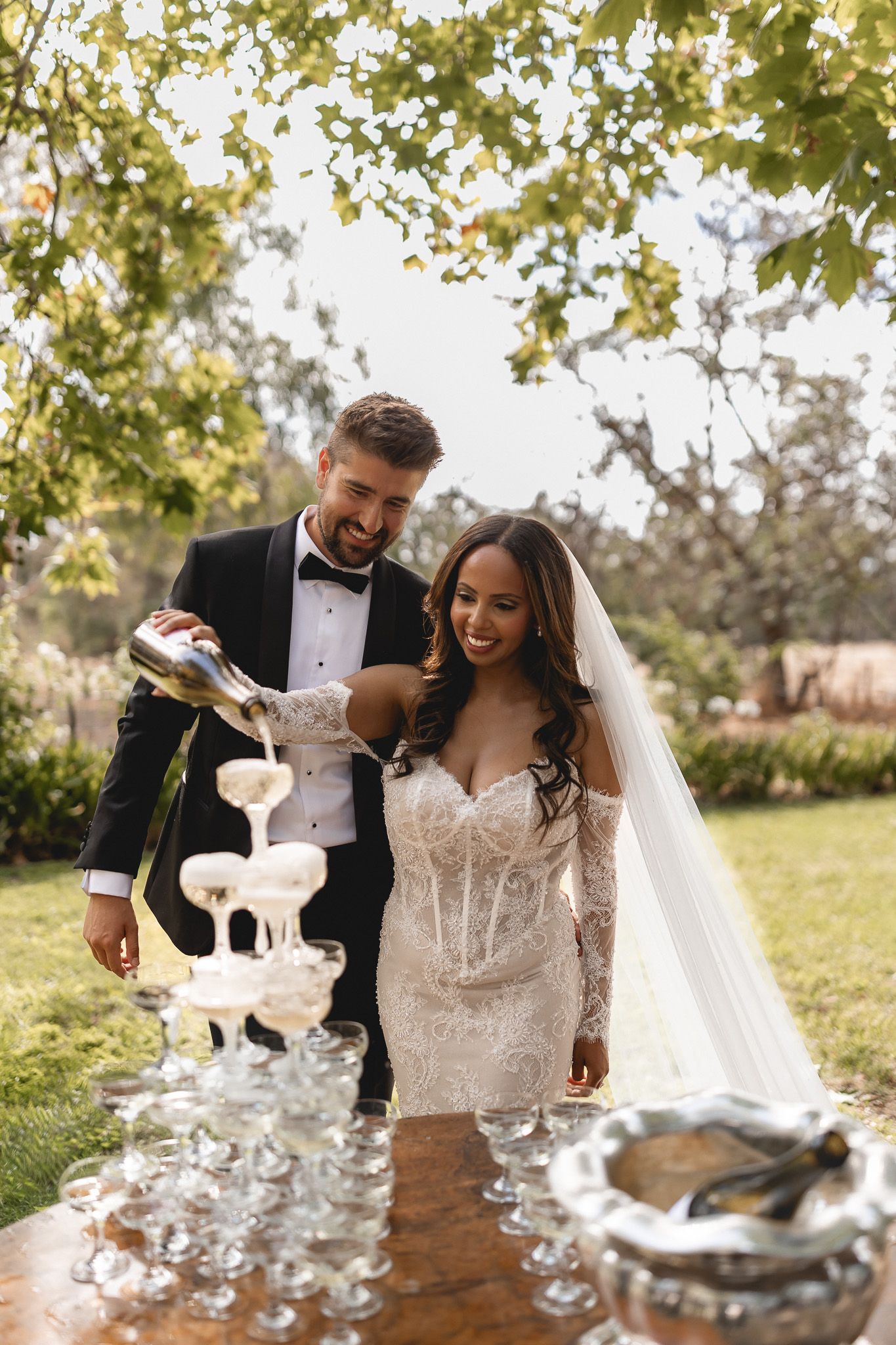Bride and groom pouring champagne tower outdoors at wedding reception under green trees.
