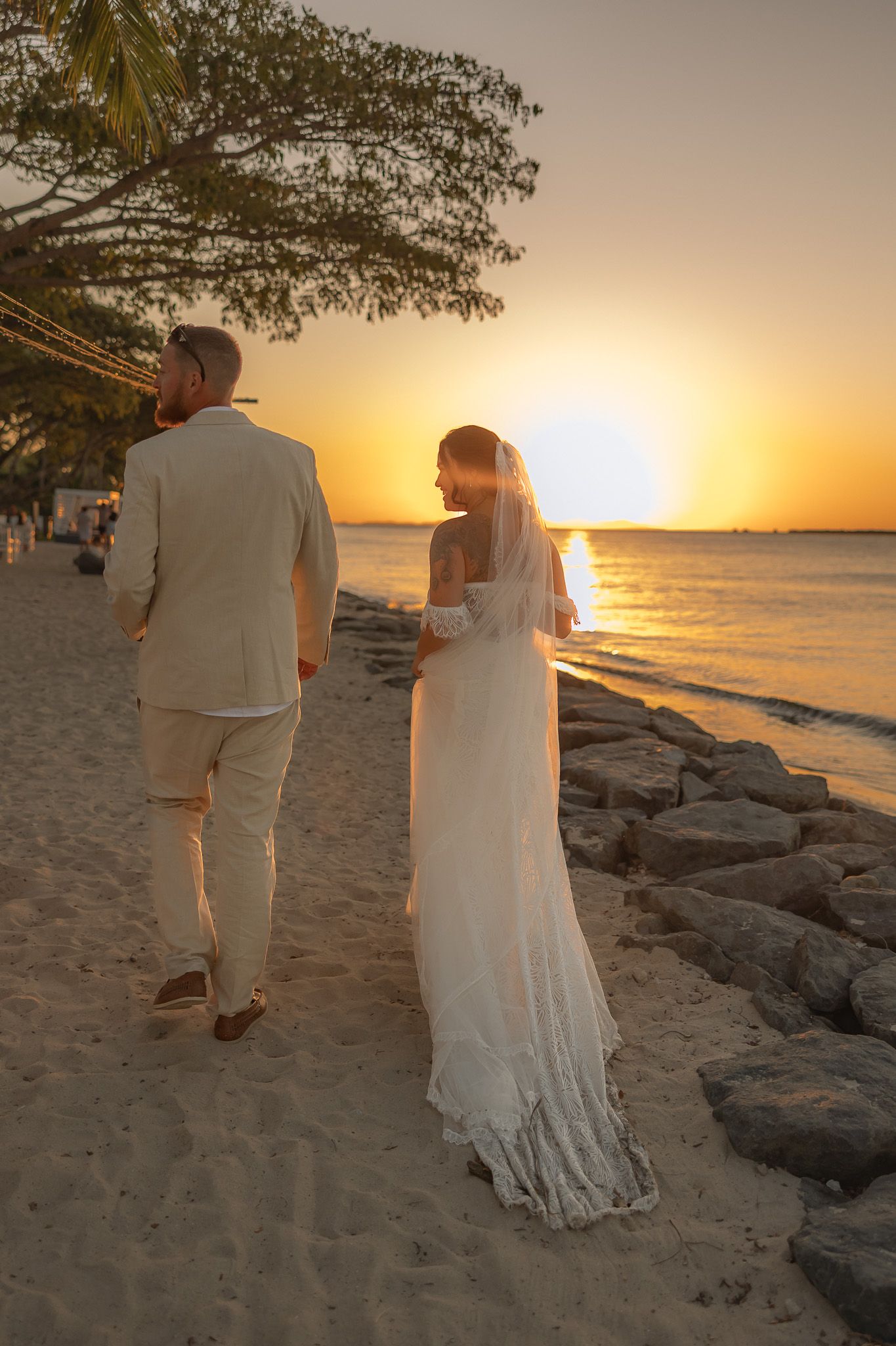 Bride and groom walk hand in hand along sandy beach at golden sunset during romantic wedding celebration.