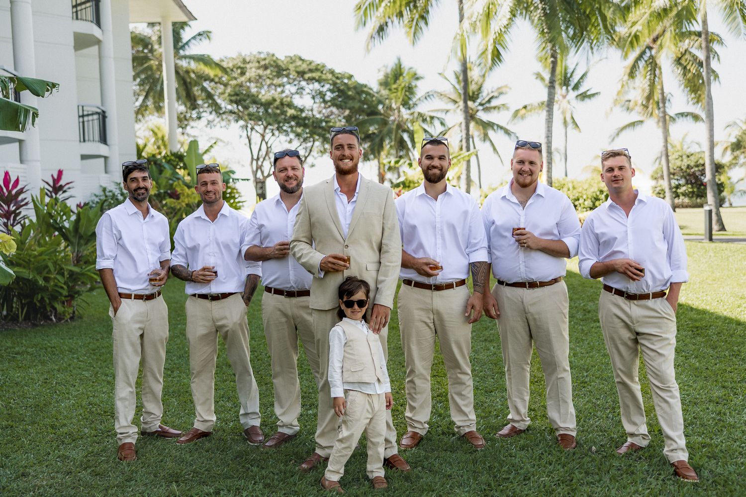 Groom and groomsmen in white shirts and khaki pants posing on green lawn with tropical palm trees backdrop.