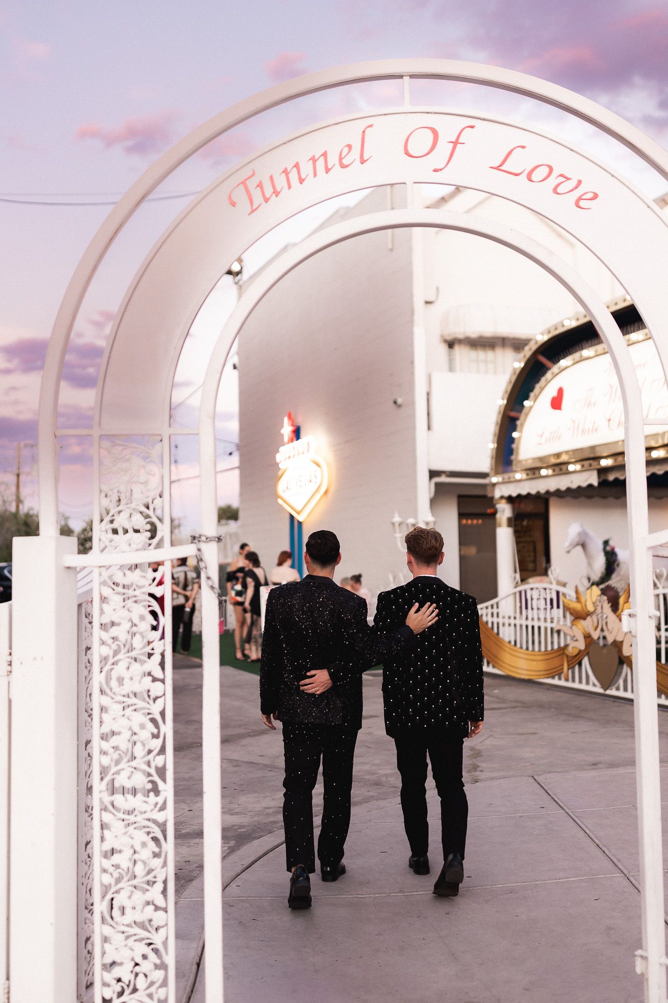Two grooms in black suits walk hand in hand through Tunnel of Love arch at Las Vegas wedding chapel.
